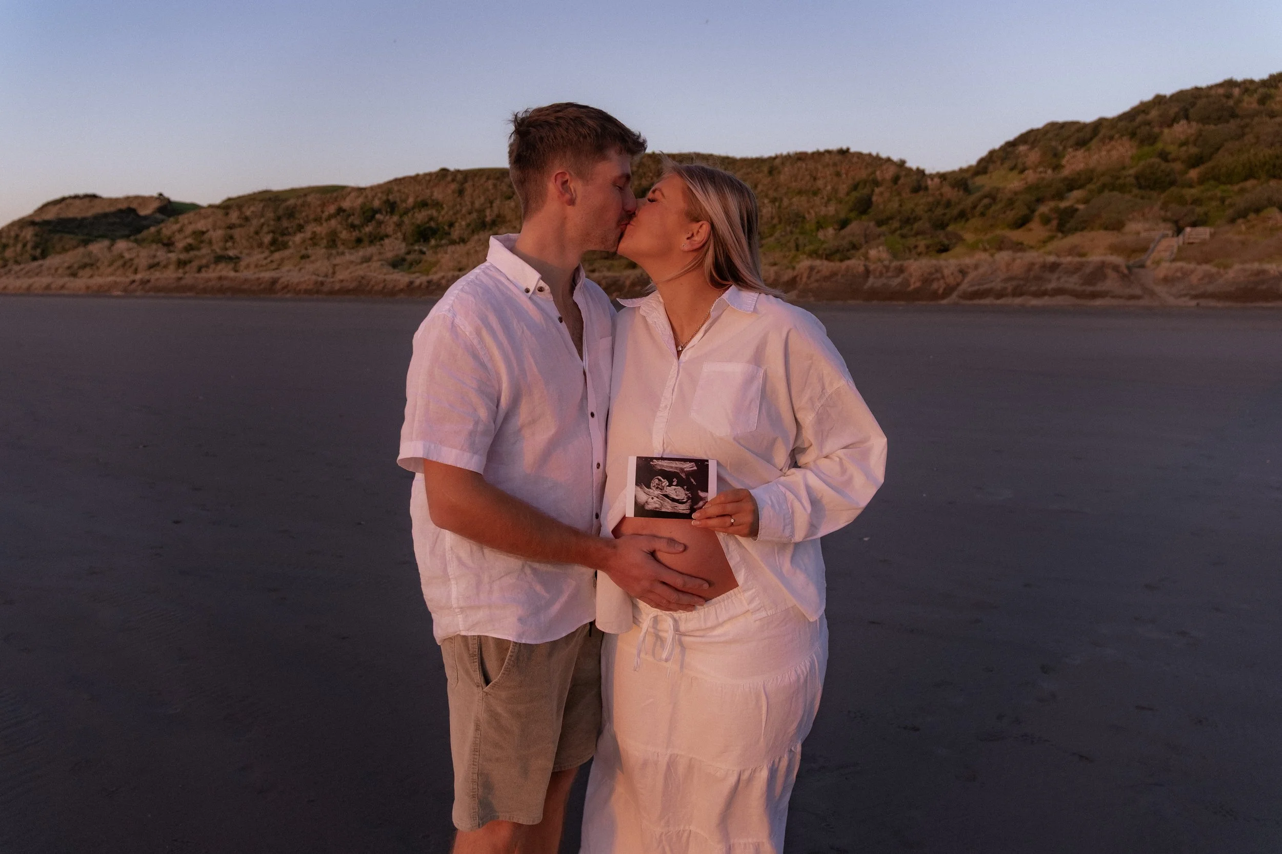 A couple on a beach at sunset, sharing a kiss, with the woman holding an ultrasound picture, suggesting pregnancy. Maternity photography. Raglan New Zealand