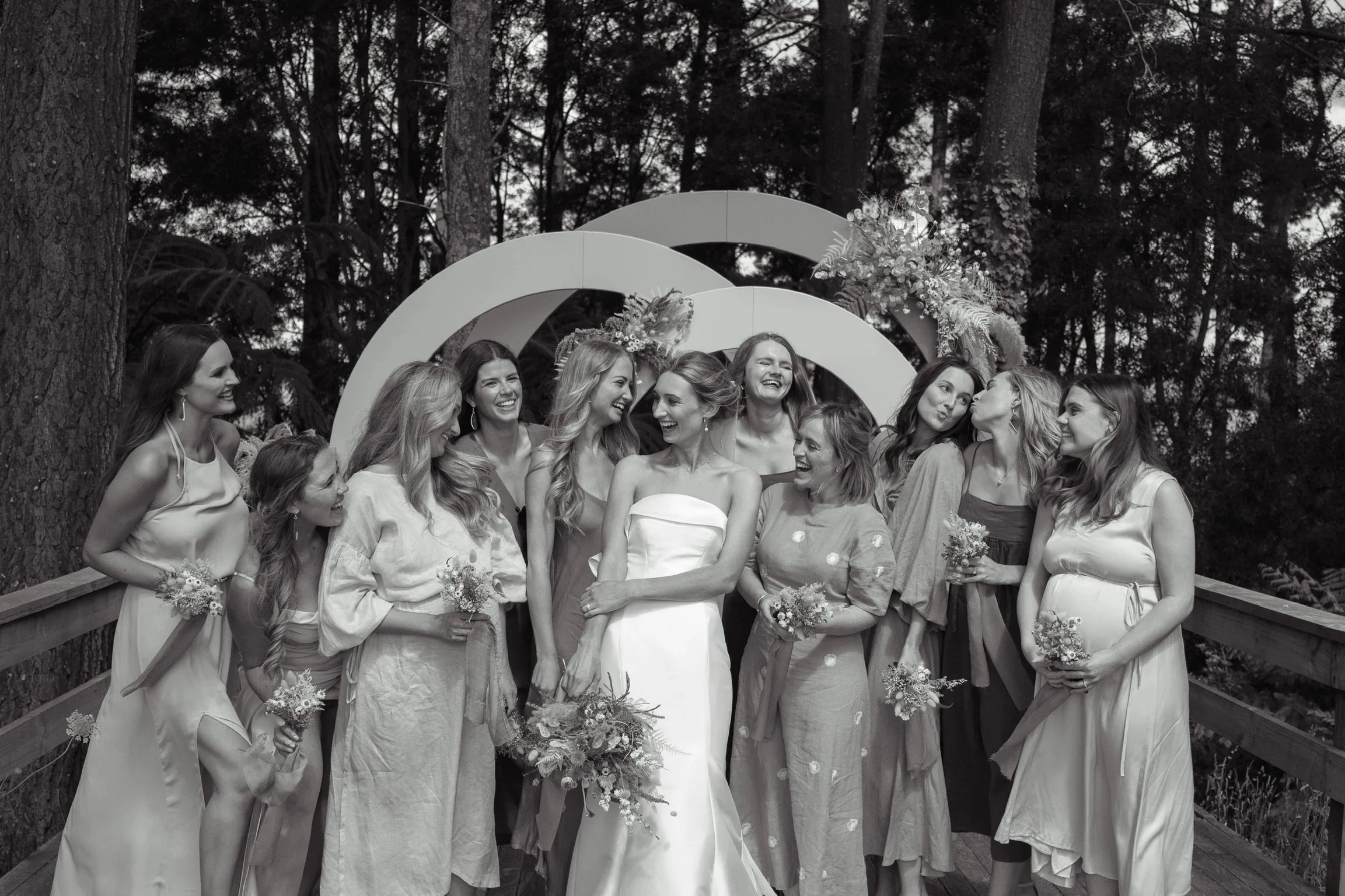 A black-and-white photo of a bride with a bouquet of flowers surrounded by her bridesmaids, standing on a wooden deck outdoors with trees in the background. The women are smiling and laughing.