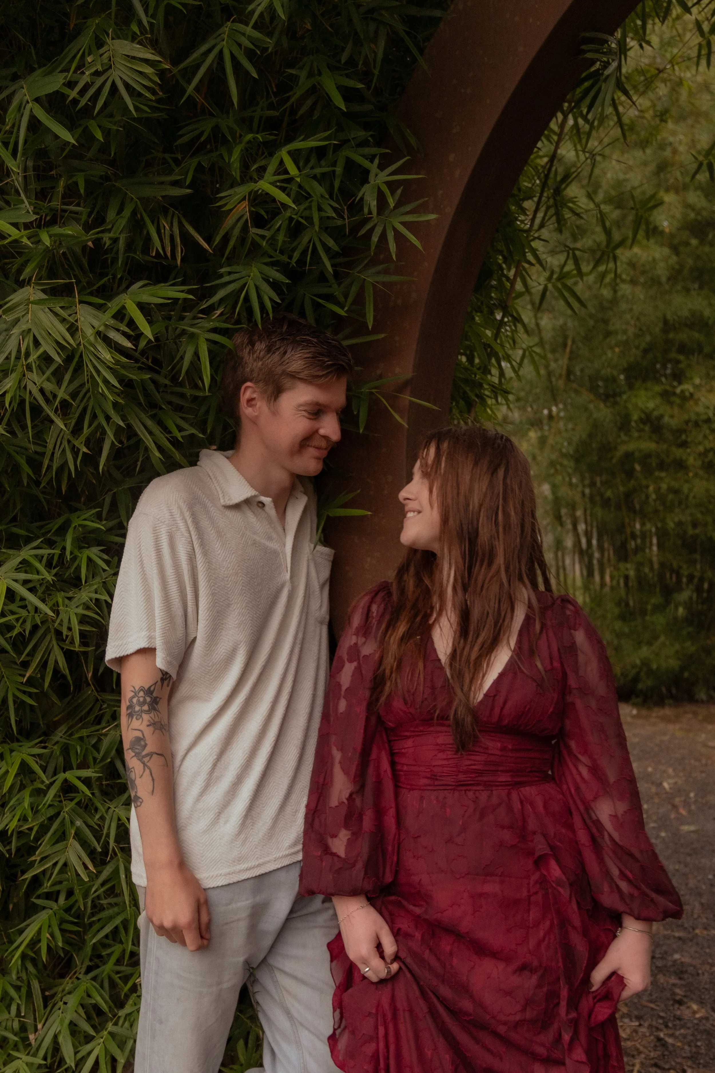 A couple standing outdoors, smiling at each other, among green foliage and trees, with the man leaning against a tree and the woman holding her dress. Couples photography Hamilton New Zealand. Engagement photography Hamilton New Zealand. 