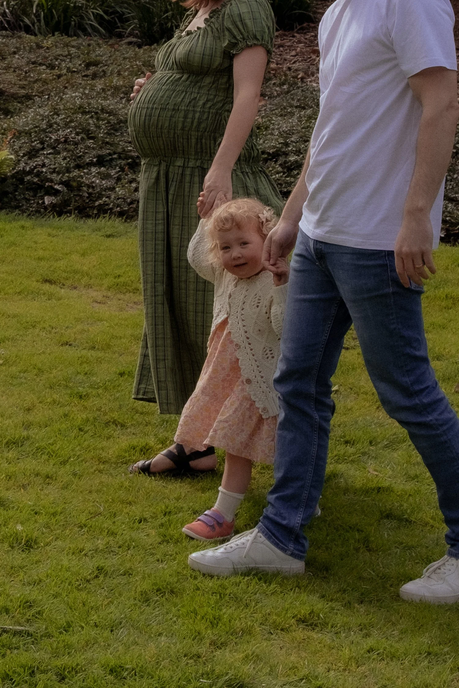 A young girl with curly hair and a pink floral dress, holding hands with a man in a white t-shirt and jeans, standing on grass alongside a pregnant woman in a green dress. The girl appears to be crying or upset. Family Photography Hamilton New Zealan