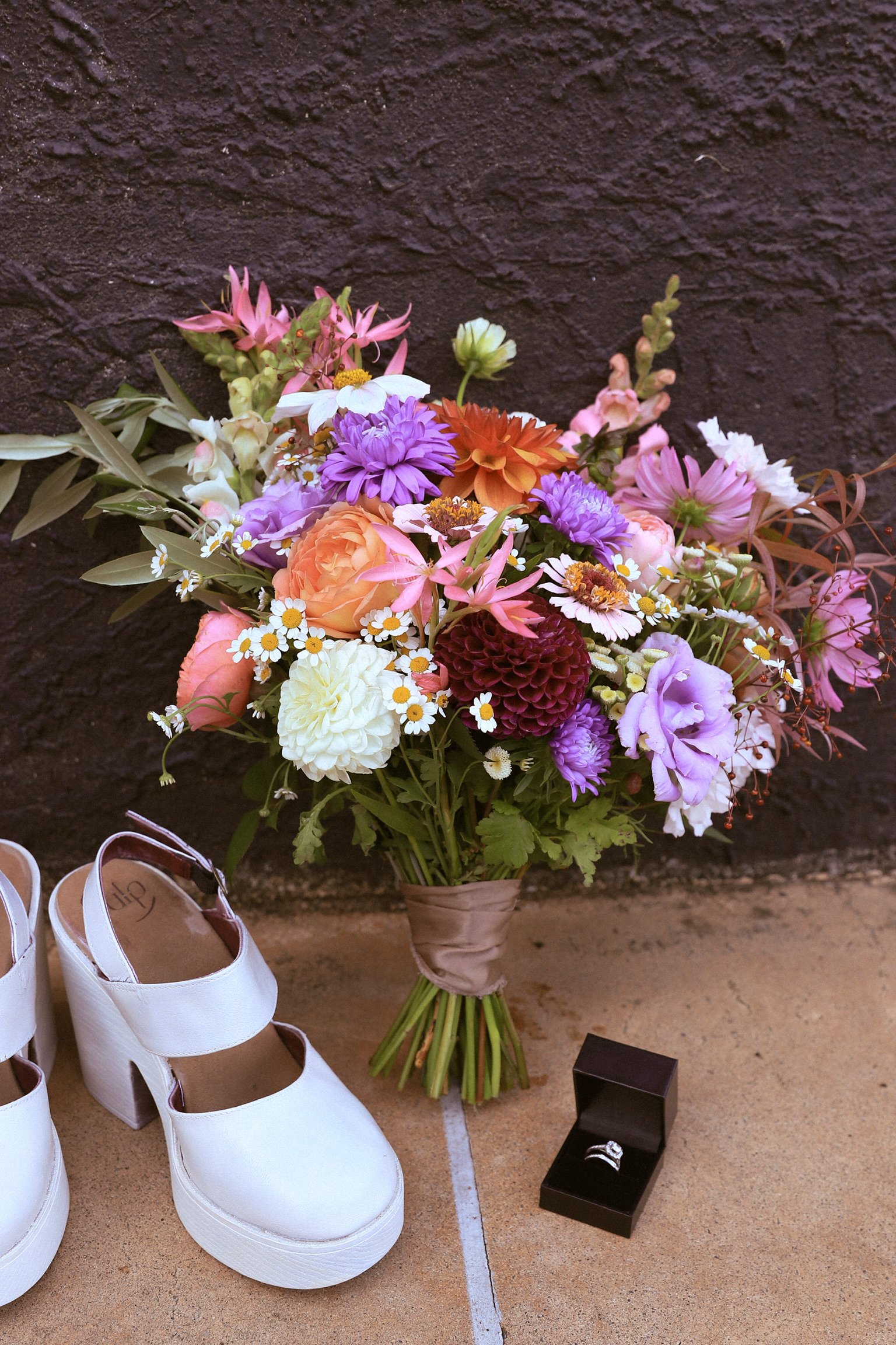 A colorful bouquet of flowers, a pair of white high-heeled shoes, and an engagement ring in a black box are placed on a floor against a dark wall. Wedding Photography Waikato New Zealand. Wedding Photography Hamilton New Zealand
