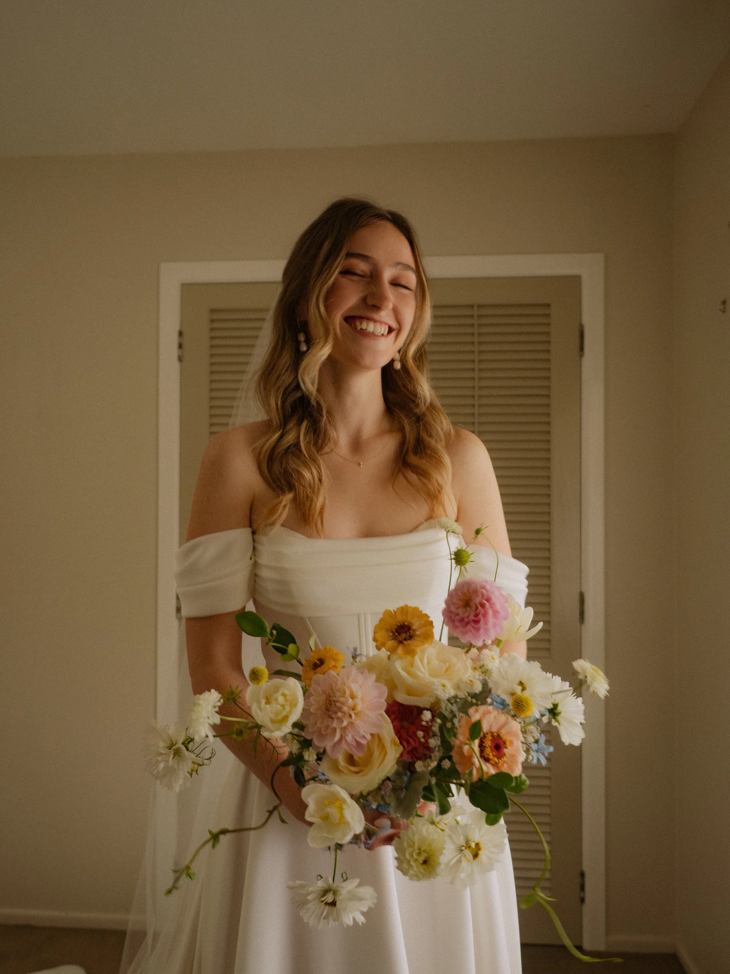 A young woman with wavy hair wearing a white off-the-shoulder dress, smiling happily while holding a large bouquet of colorful flowers including pink, yellow, and white blooms, standing in a room with a closed door and beige walls.
