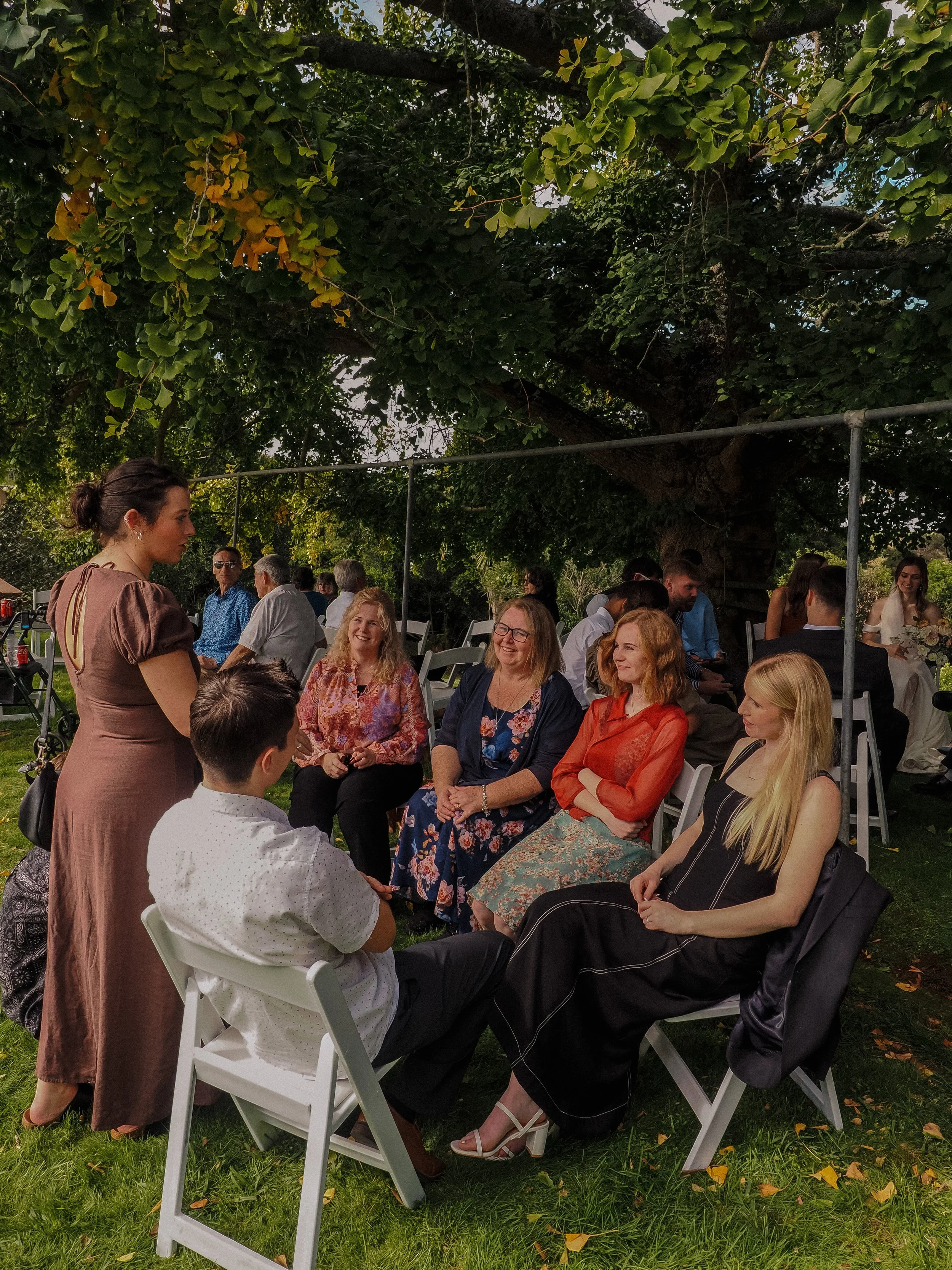 Group of people sitting and standing outdoors at a gathering or celebration under a large tree with green leaves.  Wedding Photography Waikato New Zealand. Wedding Photography Hamilton New Zealand. Northland Wedding Photography