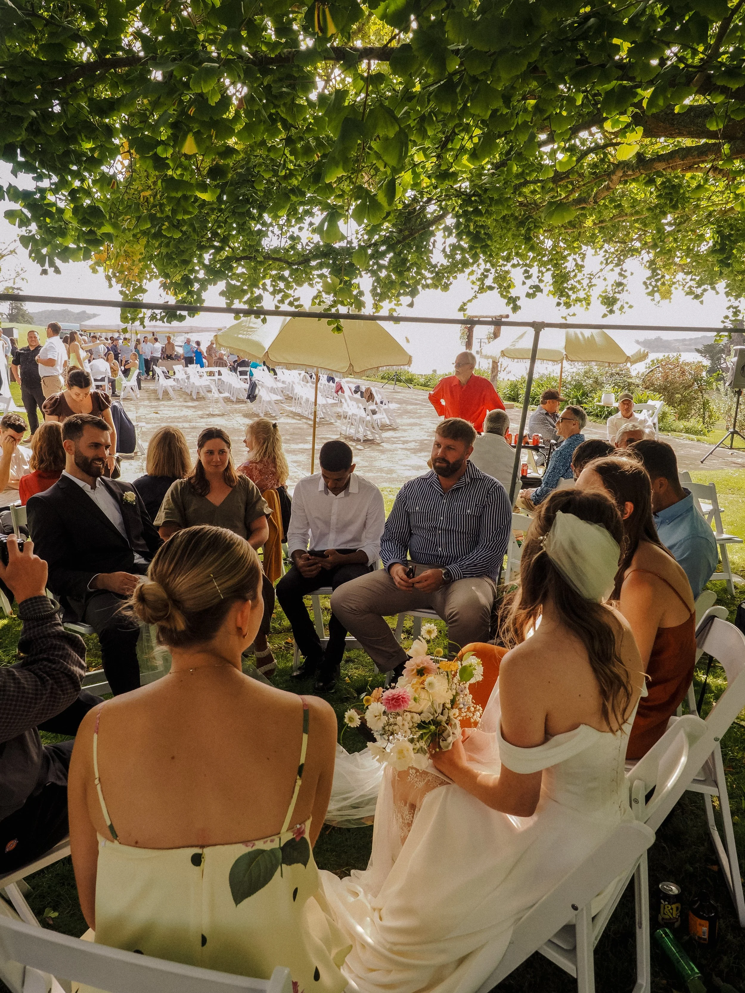 People at an outdoor wedding ceremony, sitting under a tree, with the bride holding a bouquet, and guests seated on white chairs, some using sunglasses, during daytime.  Wedding Photography Waikato New Zealand. Wedding Photography Hamilton 