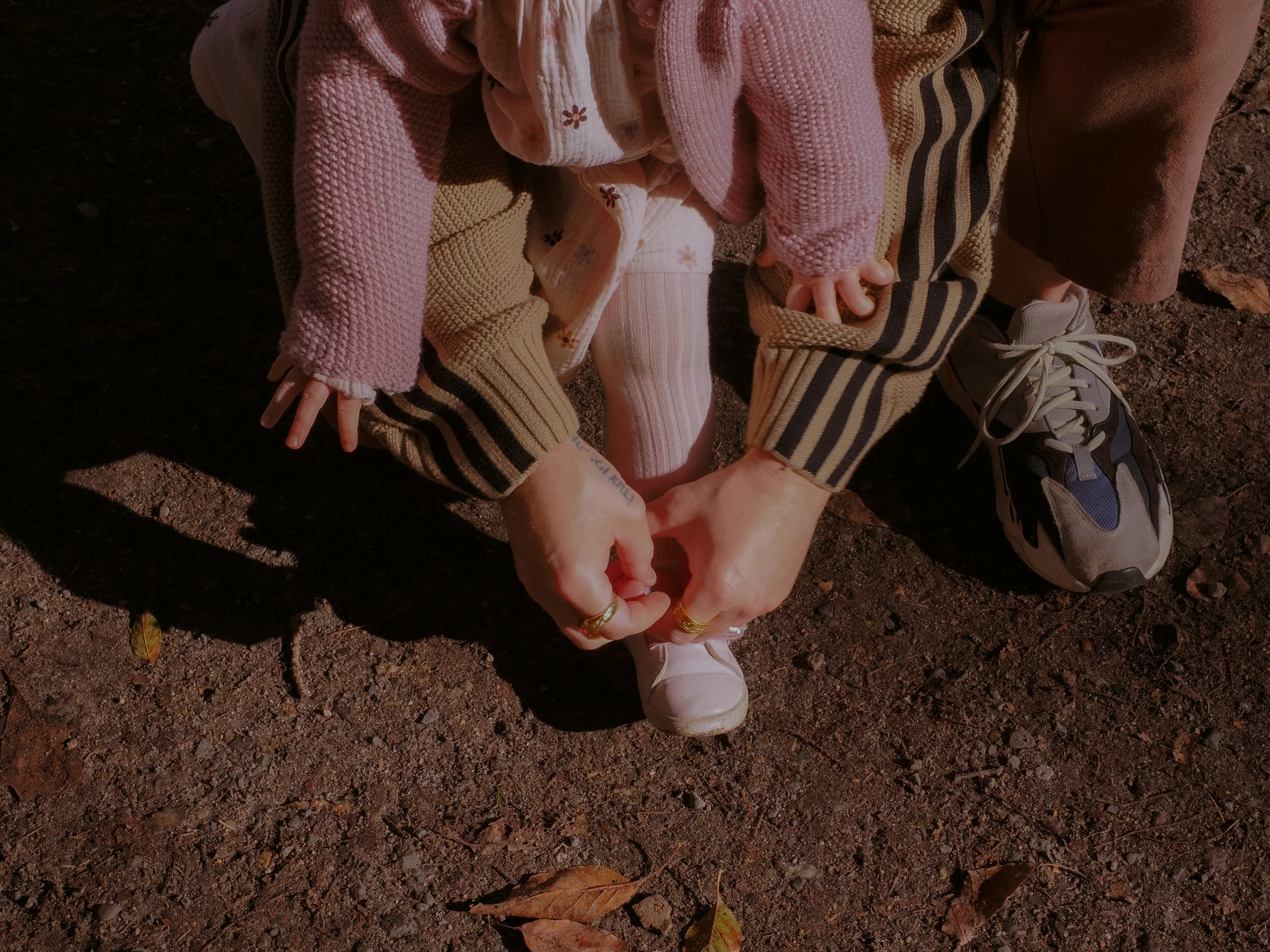 A child tying their shoelace while an adult helps, both kneeling on dirt ground with fallen leaves. Family Photography Waikato New Zealand. Family Photography Hamilton New Zealand. Northland Wedding Photography