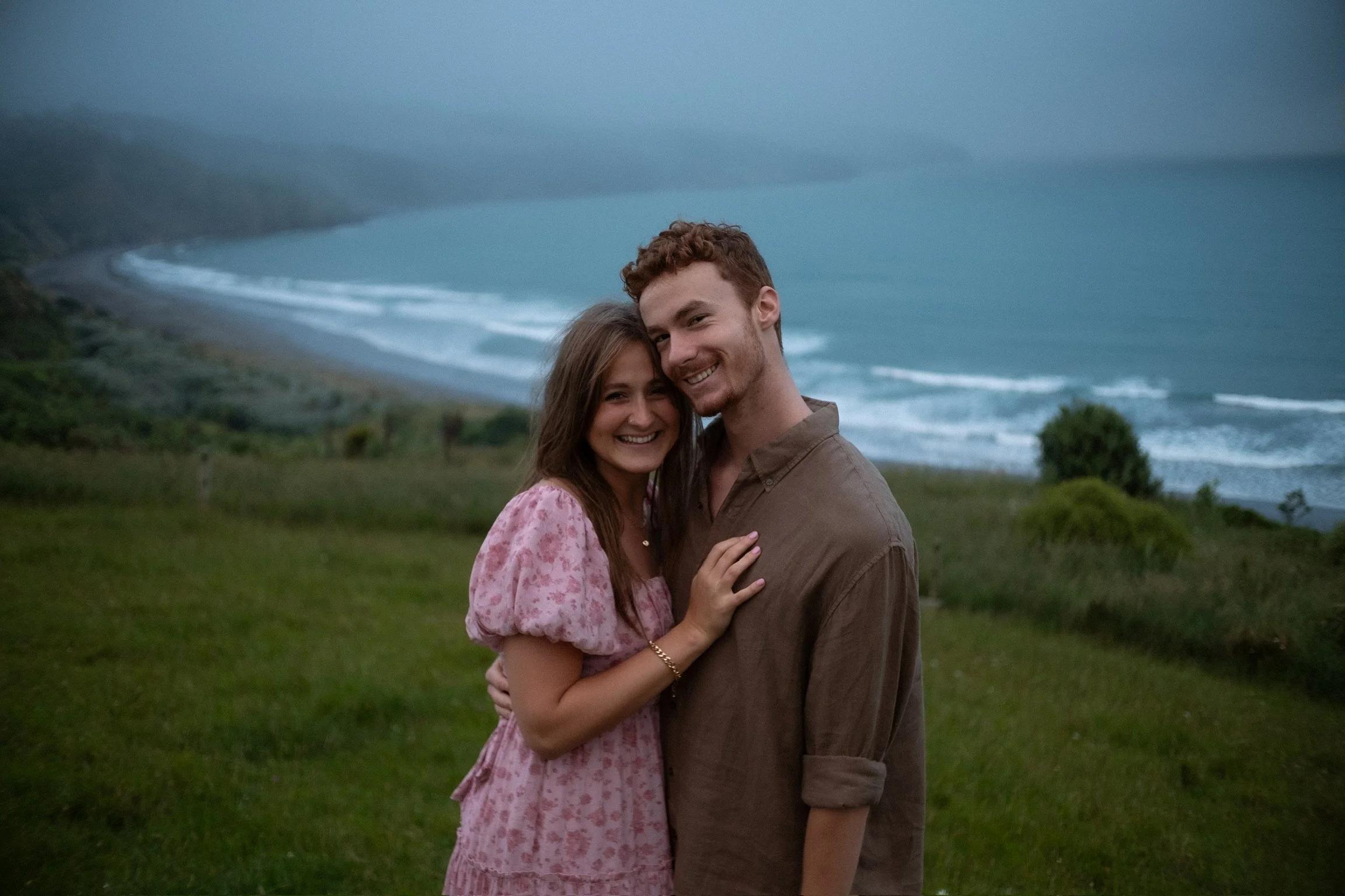 A smiling couple standing close together on a grassy hillside, with an ocean and foggy coastline in the background. Couples photography Raglan New Zealand. Engagement Photography Hamilton New Zealand. Whangarei Photographer