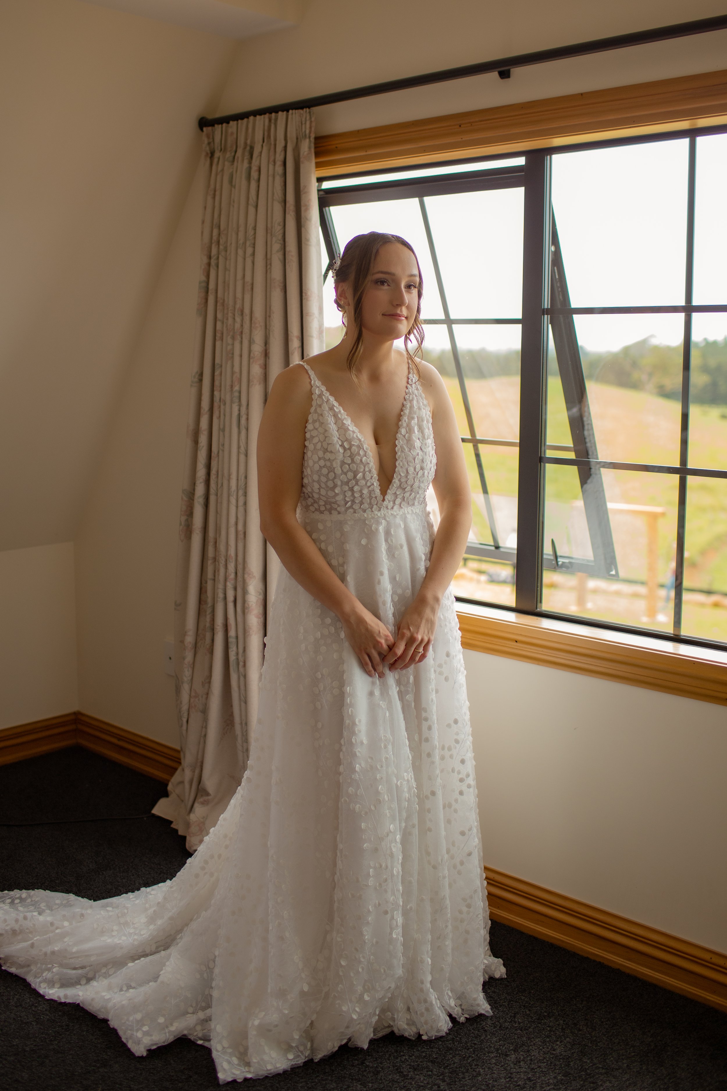 A bride in a white wedding dress stands by a window in a room with beige walls and wooden trim, looking outside with a serene expression.  Wedding Photography Waikato New Zealand. Wedding Photography Hamilton New Zealand. Northland Wedding Photo