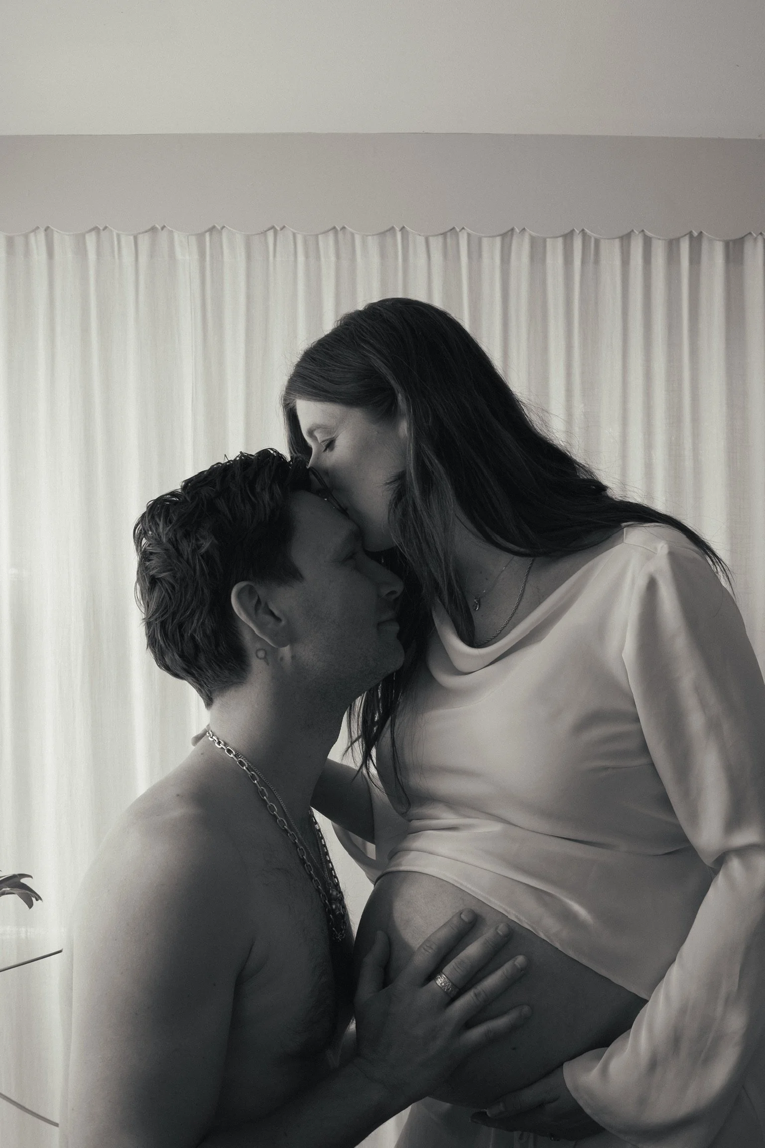 A black and white photo of a pregnant woman and a man. The woman is leaning forward to kiss the man on his forehead, and he is touching her pregnant belly with his hand. They are embracing intimately in front of a light-colored curtain. Maternity