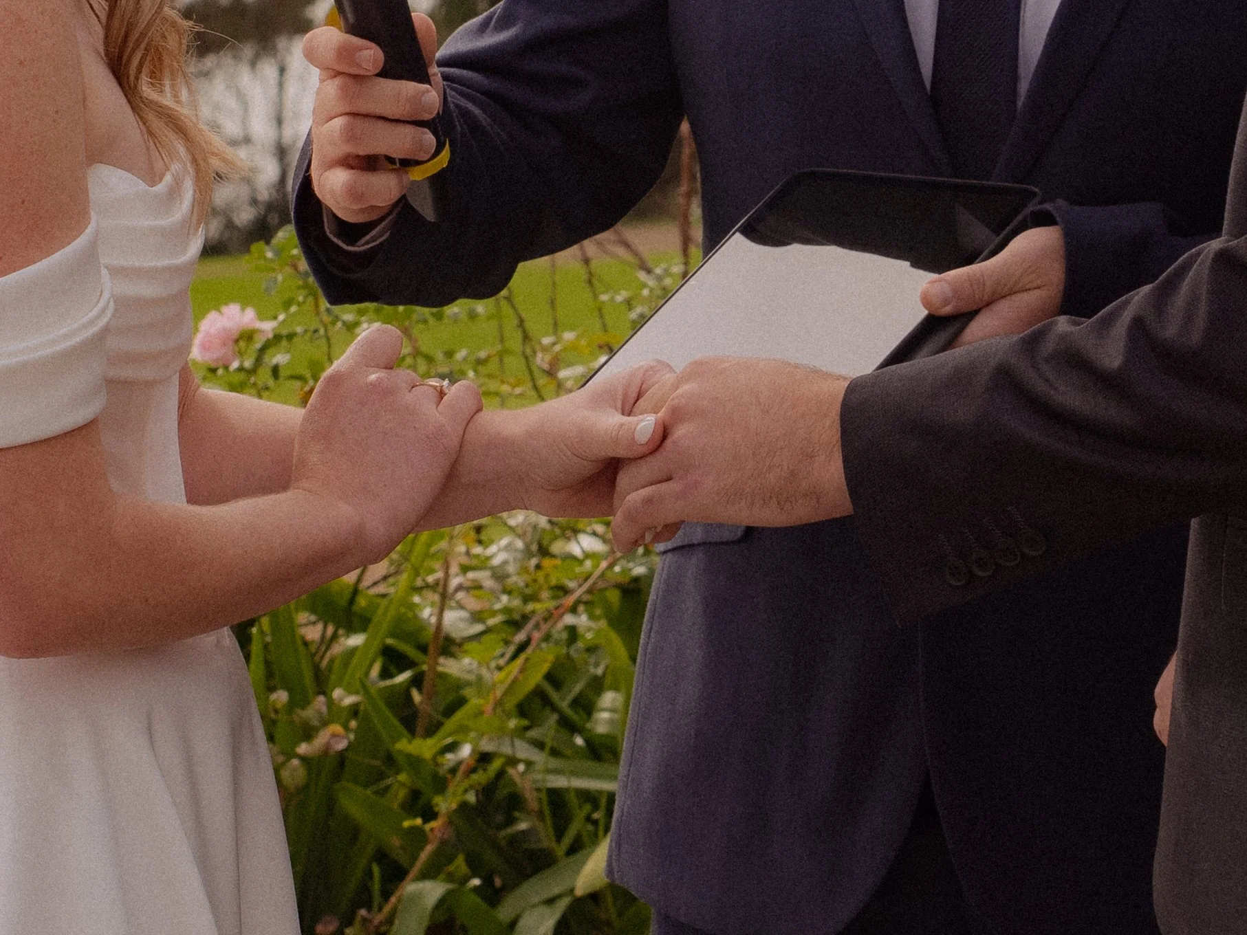 A couple exchanging wedding vows, holding hands, with an officiant present, outdoors with greenery and flowers in the background.  Wedding Photography Waikato New Zealand. Wedding Photography Hamilton New Zealand. Northland Wedding Photography