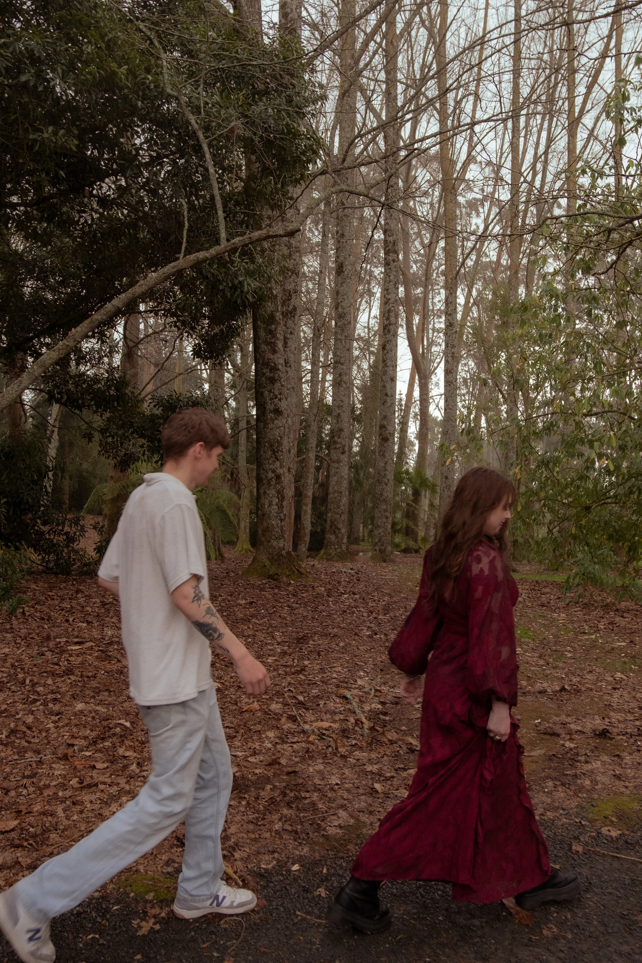 A young man and woman walking through a wooded forest trail during daytime. The man has short brown hair, light-colored clothing, and visible tattoos on his left arm. The woman has long brown hair and is wearing a long, dark red dress and black boots