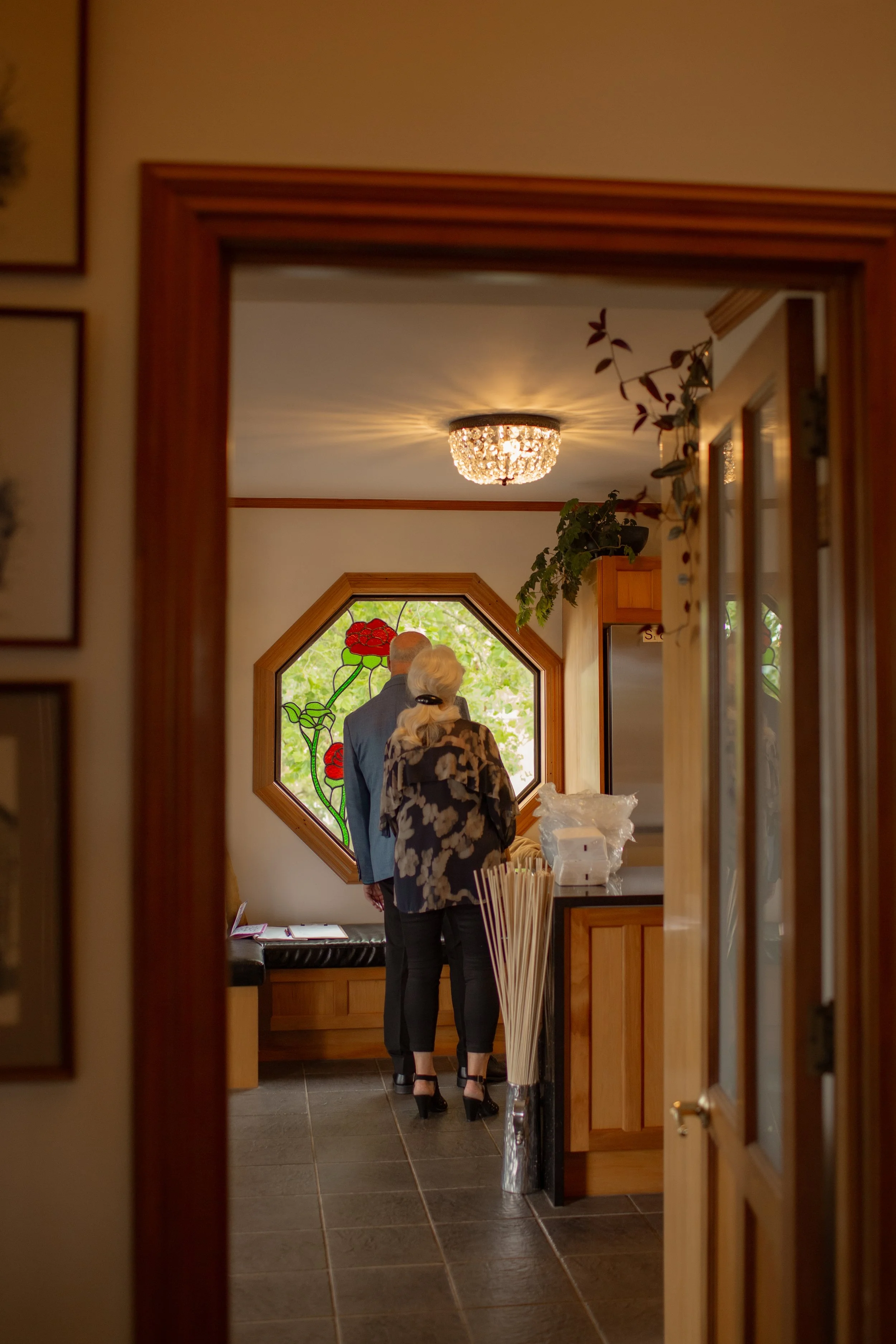Two people, an older man and woman, are standing inside a room, facing a stained-glass window with red flowers. The photo is taken through a doorway, showing a chandelier on the ceiling, a refrigerator, and some plants.