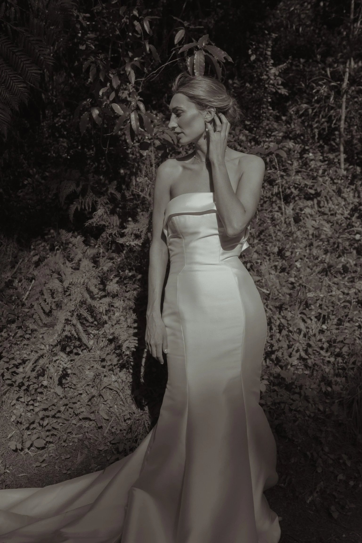 A woman in an elegant strapless satin gown standing amidst a lush outdoor backdrop with dense foliage, her hand gently touching her neck.  Wedding Photography Waikato New Zealand. Wedding Photography Hamilton New Zealand. Northland Wedding Photograph