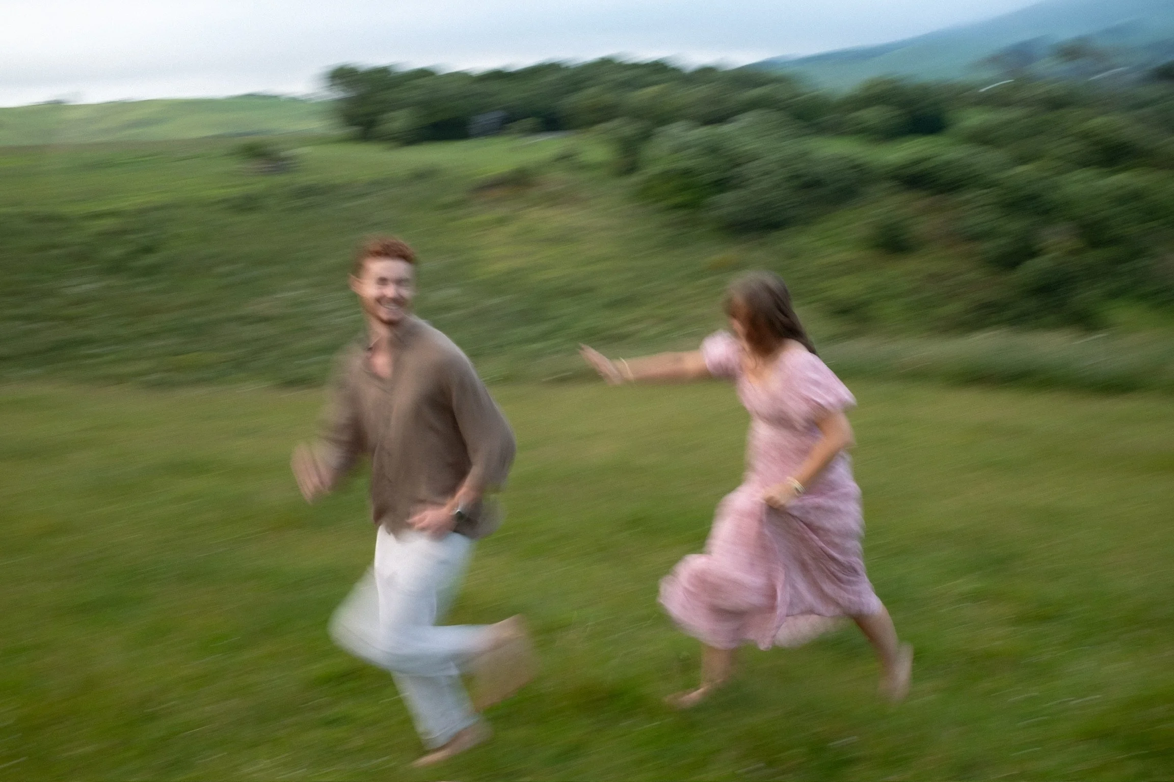 A man and woman running outdoors across a grassy field, with the woman reaching out towards the man. The background includes rolling hills and some trees, and the photo appears blurred due to motion. Couples photography Raglan New Zealand. Engagement