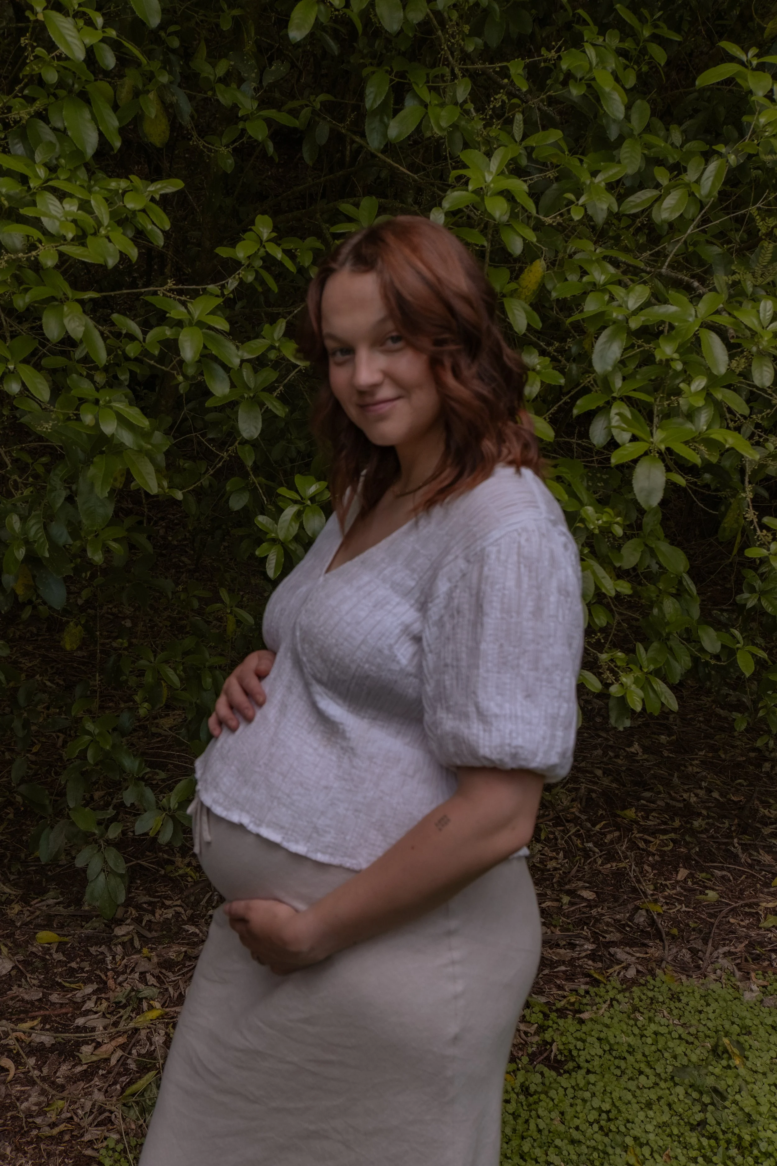 A pregnant woman with shoulder-length red hair standing outdoors in front of green bushes, wearing a white blouse and beige skirt, smiling softly. Maternity Photography, Hamilton New Zealand Photographer