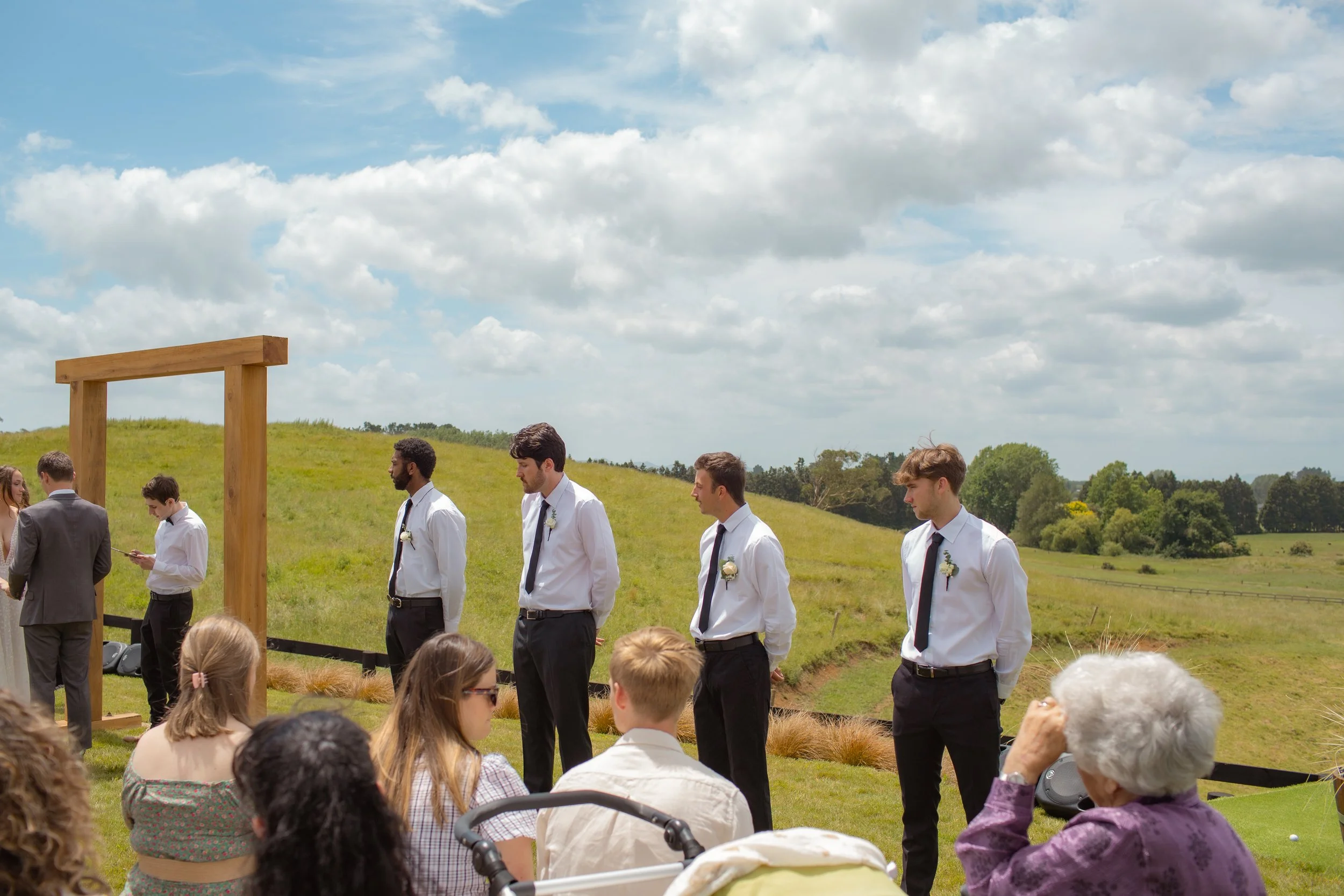 A wedding ceremony outdoors with groomsmen standing in a line on a grassy field, dressed in white shirts, black pants, and black ties, against a backdrop of rolling green hills and a partly cloudy sky. Guests sit in the foreground, including an elder