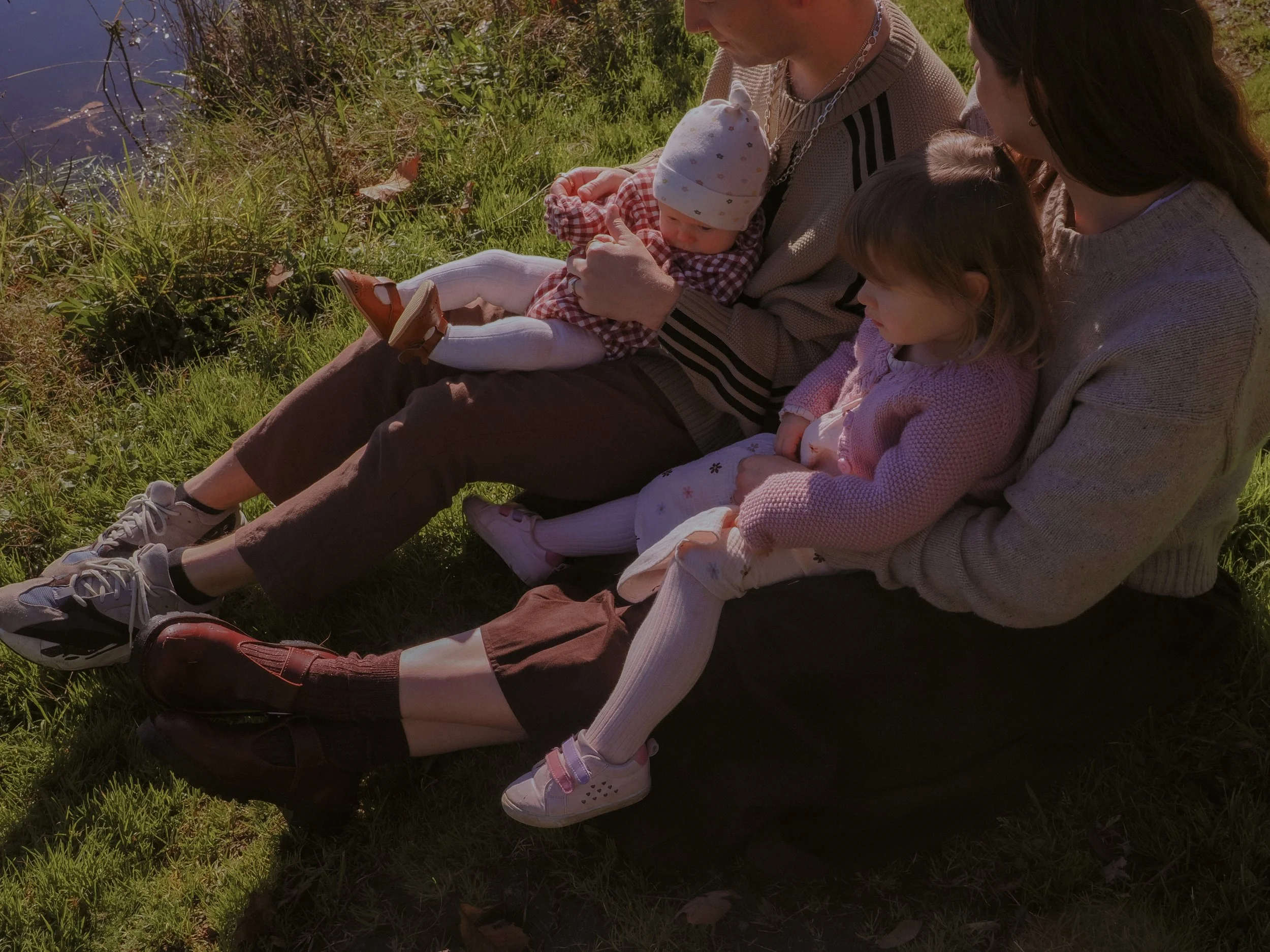 A family sitting on the grass by a lakeside during daytime, with a baby, a young girl, and two adults. Family Photography Waikato New Zealand. Family Photography Hamilton New Zealand. Northland Wedding Photography
