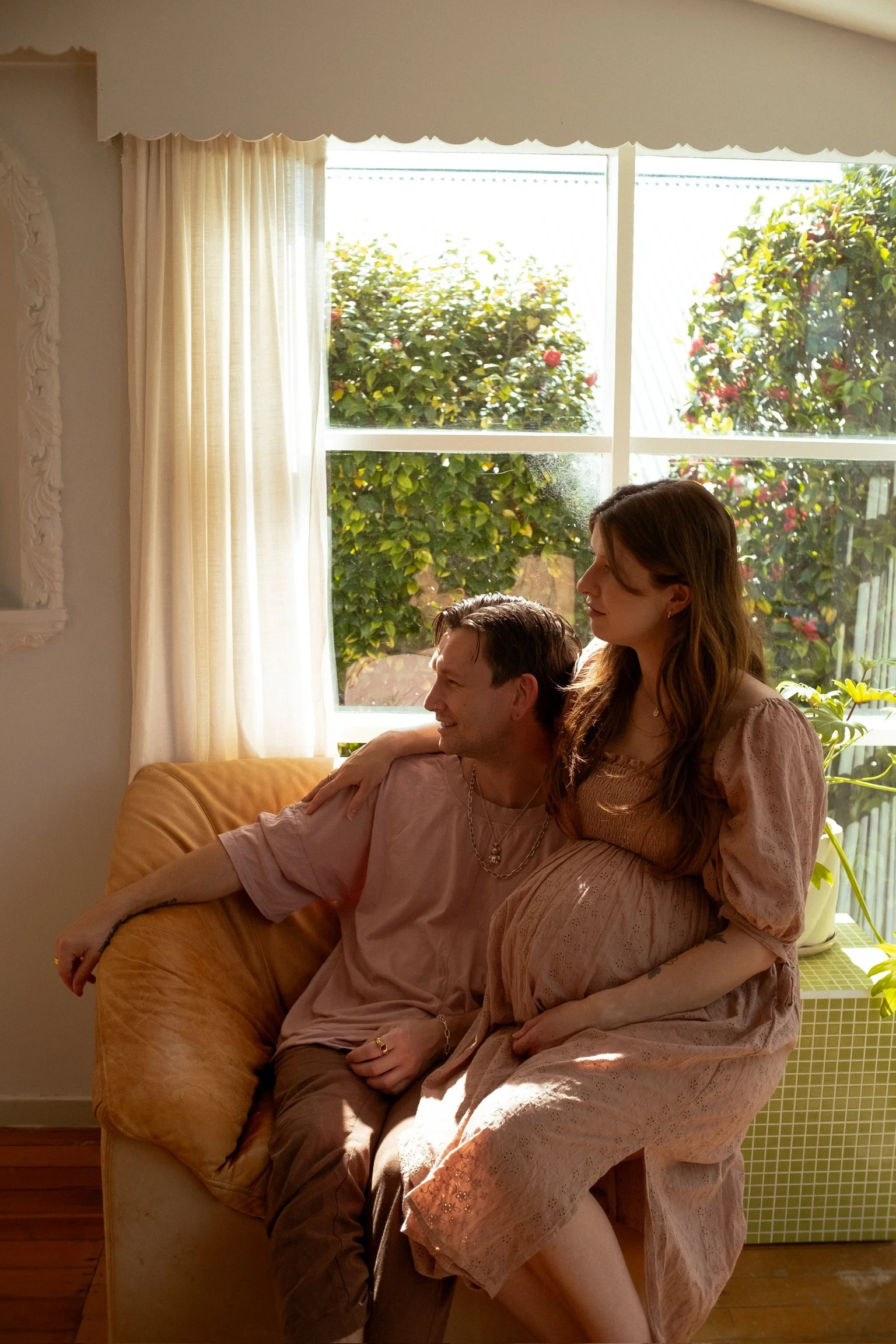A man and a pregnant woman sitting on a beige sofa in a sunlit room with a large window and green outdoor plants. The man has his arm around the woman's shoulders, and they are smiling and looking into each other's eyes. Maternity Photographer