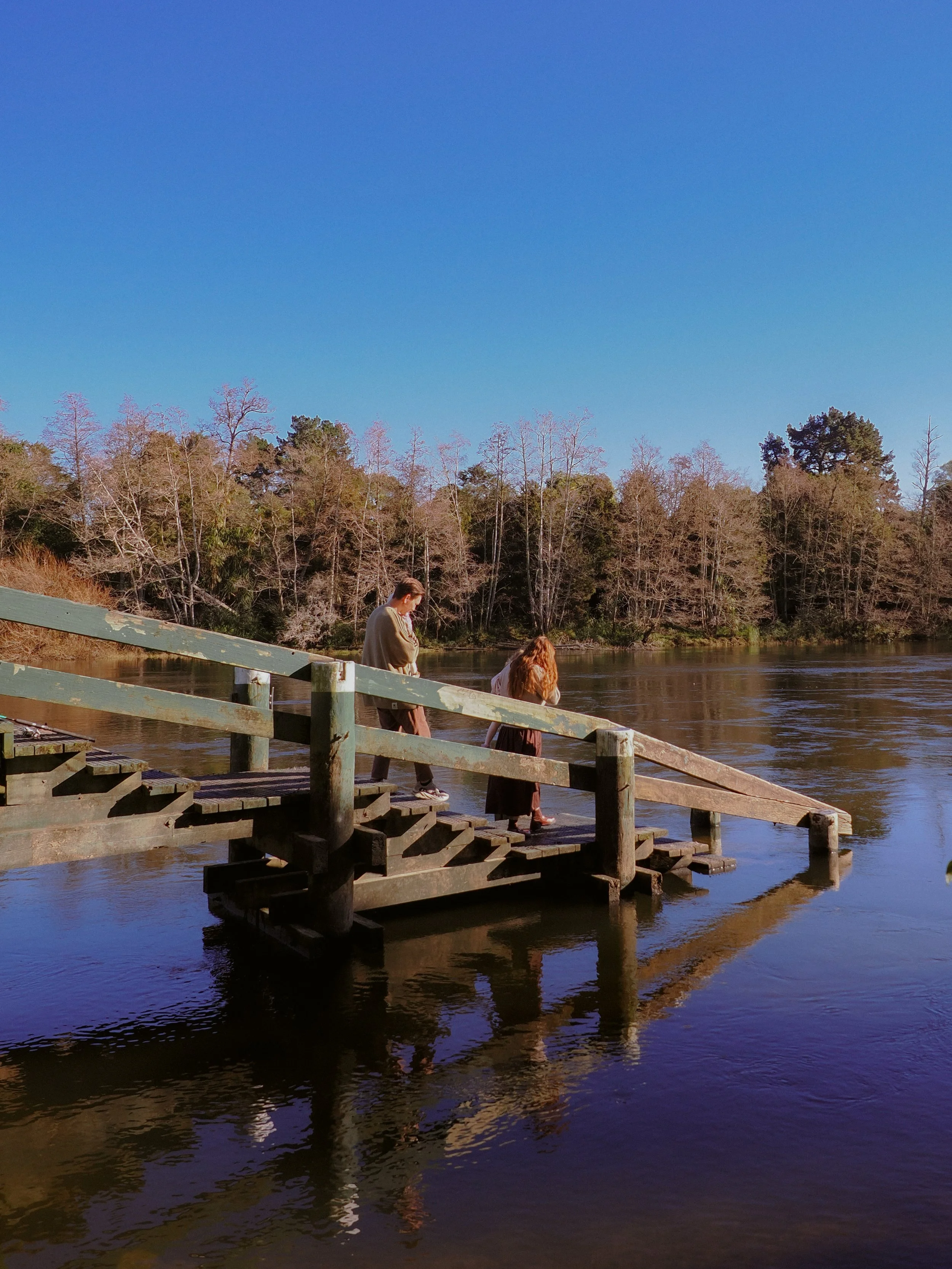 Two women walking down a wooden dock into a river, with trees on the far bank and a clear blue sky above. Family Photography Waikato New Zealand. Family Photography Hamilton New Zealand. Northland Wedding Photography