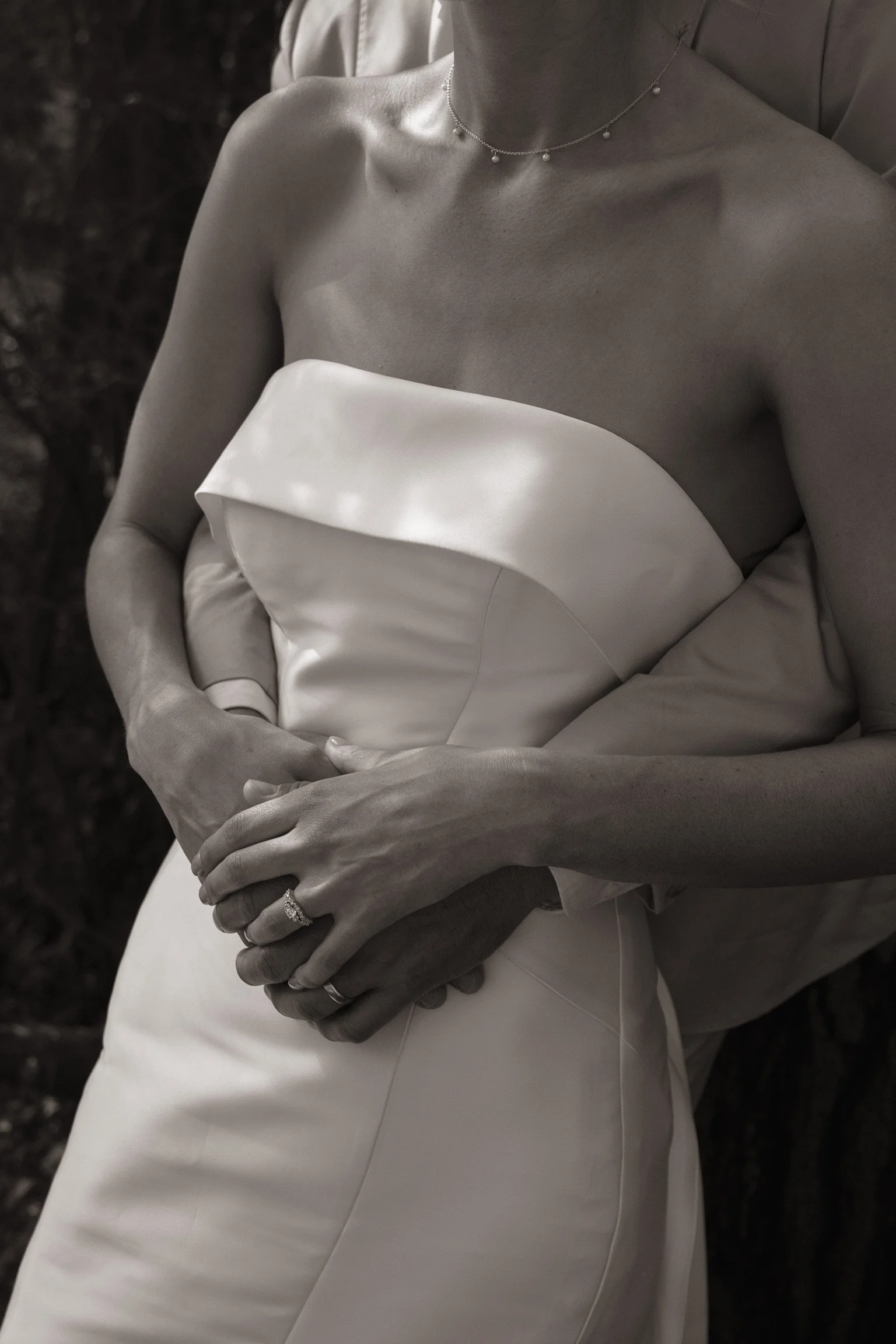 A bride wearing a strapless satin wedding gown, holding her hands over her midsection, with an engagement ring and wedding band visible on her fingers, and a delicate necklace around her neck.  Wedding Photography Waikato New Zealand. Wedding Photo