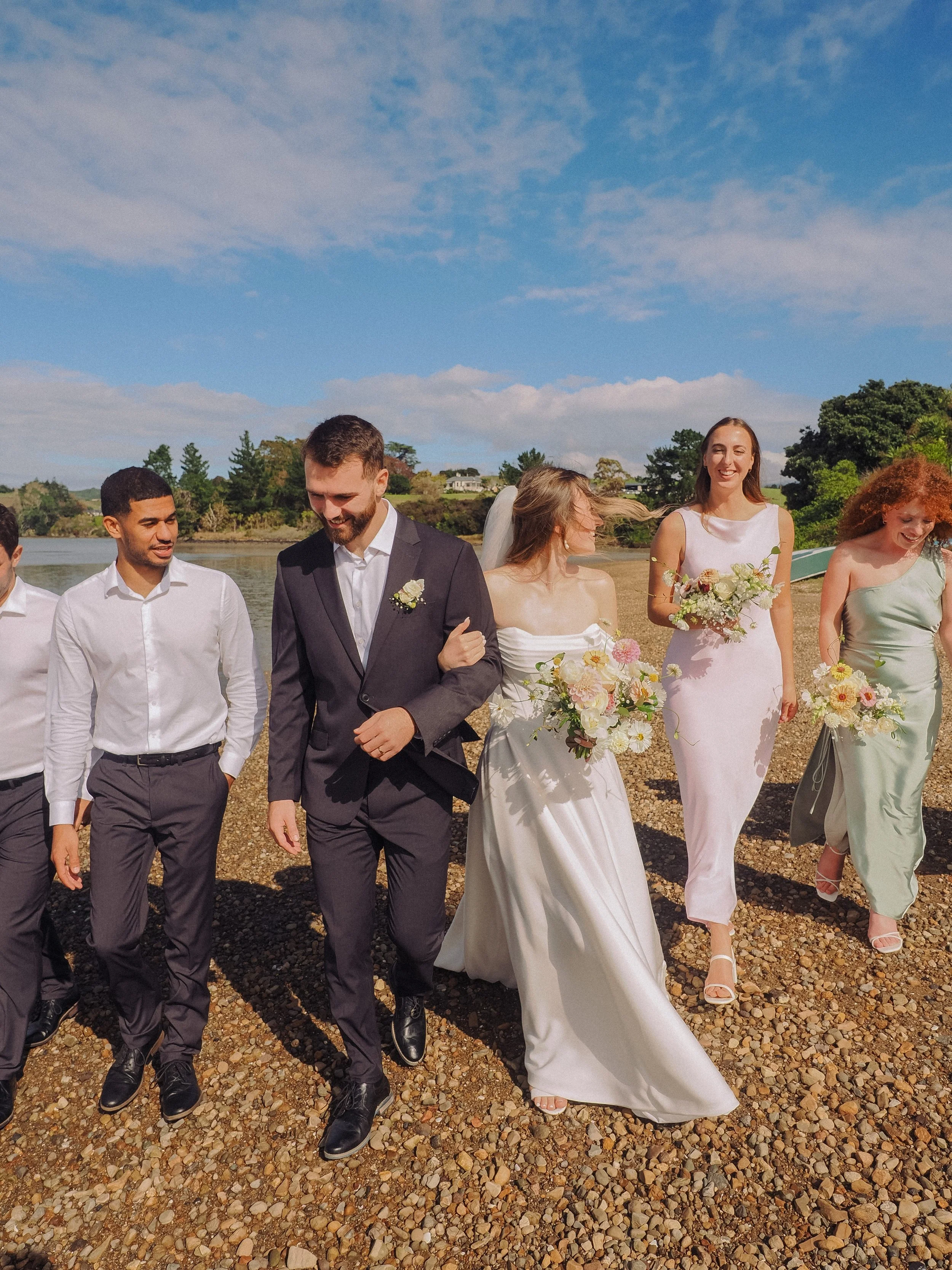 Group of people at a wedding outdoors, walking along a pebble beach with a river and trees in the background, under a partly cloudy blue sky.  Wedding Photography Waikato New Zealand. Wedding Photography Hamilton New Zealand. Northland Wedding Photo