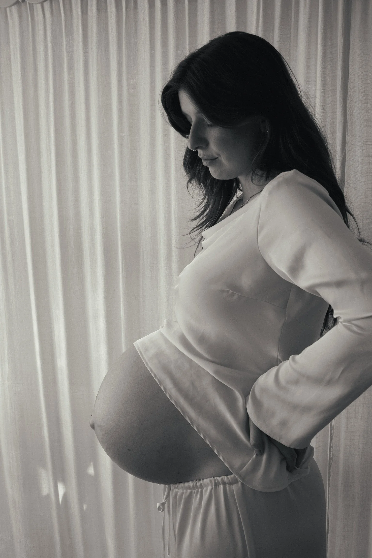 Black and white photo of a pregnant woman in a satin pajama top and pants standing sideways with hands on her back, smiling softly, with drawn curtains in the background. Maternity Photography. Hamilton New Zealand Photographer