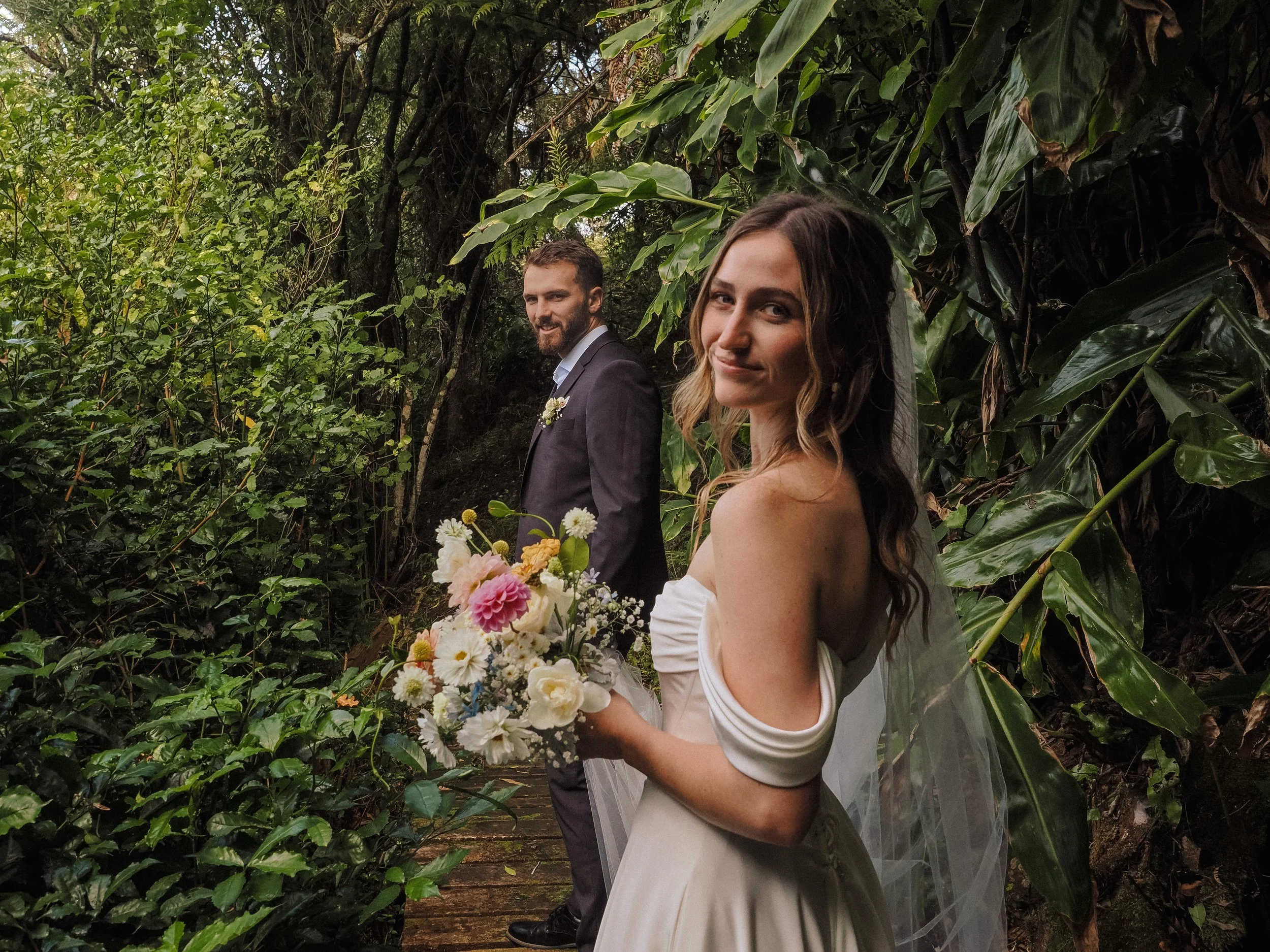 A bride and groom standing in a lush, green forest. The bride is holding a bouquet of white, yellow, and pink flowers, and is wearing a white wedding dress with a veil. The groom is dressed in a dark suit with a white shirt and a boutonniere. Both are smiling and looking at the camera.