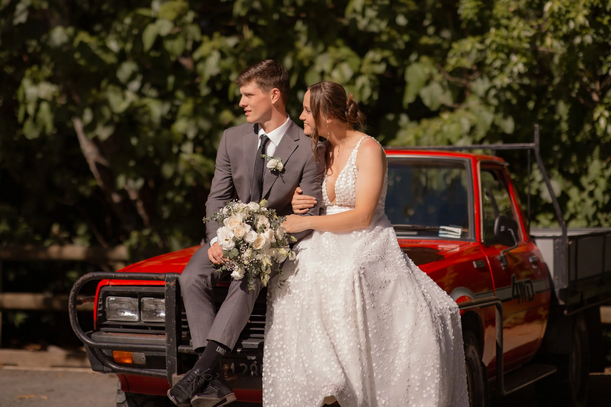 A bride and groom sitting on the hood of a red pickup truck during their wedding, with greenery in the background.  Wedding Photography Waikato New Zealand. Wedding Photography Hamilton New Zealand. Northland Wedding Photography