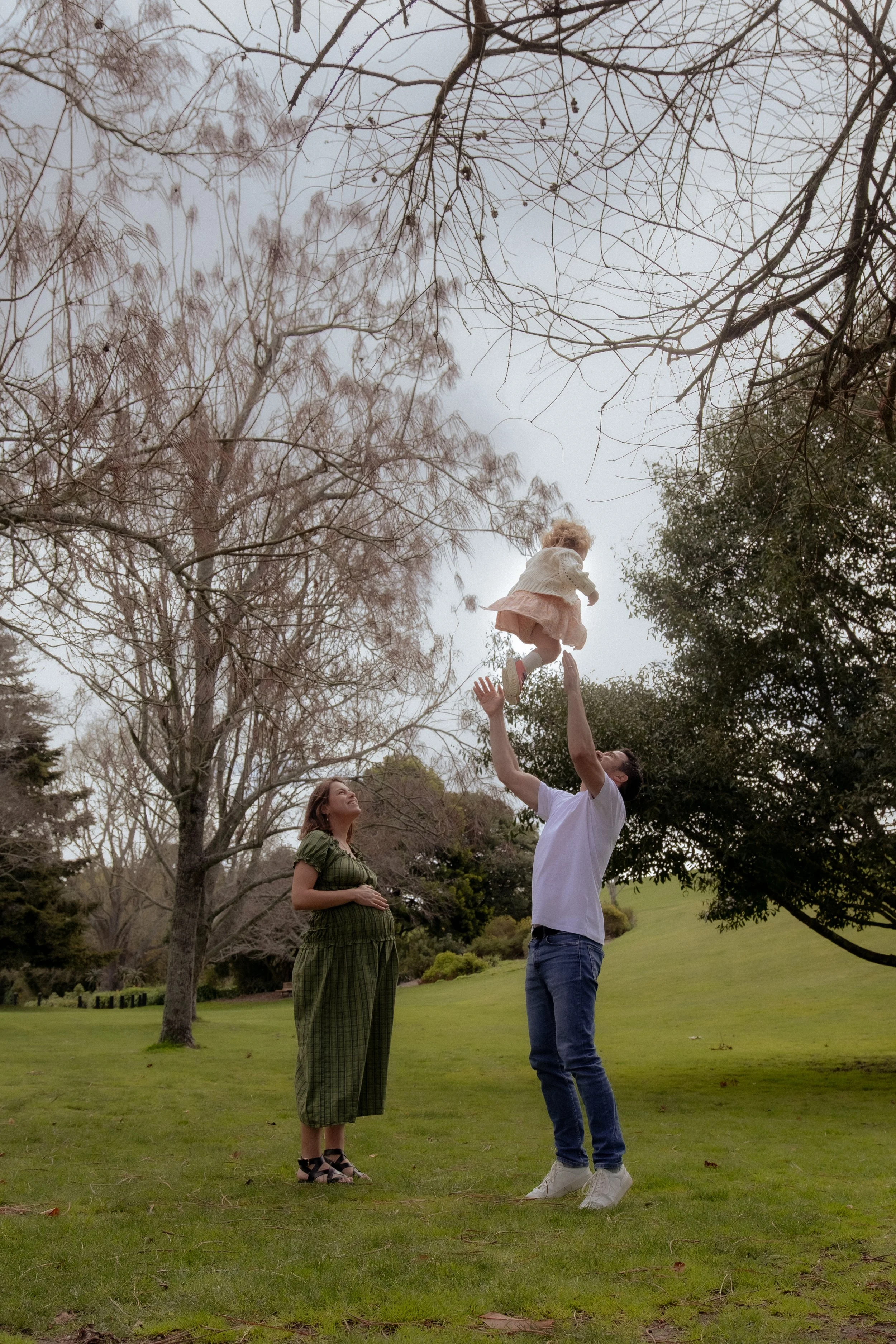 Family playing in a park during daytime with a man lifting a girl in the air and a pregnant woman watching, surrounded by trees and green grass. Family photography, maternity photography Hamilton New Zealand