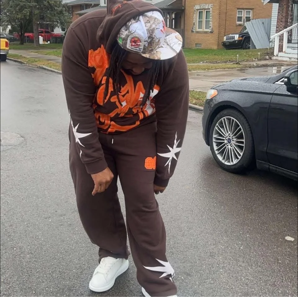 Person wearing a brown tracksuit with orange and white designs, standing on a street next to a car.