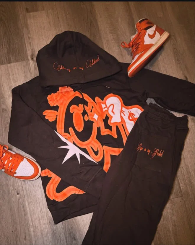 Fashion ensemble with a black hoodie featuring orange graffiti-style graphics, black joggers, and orange and white sneakers.