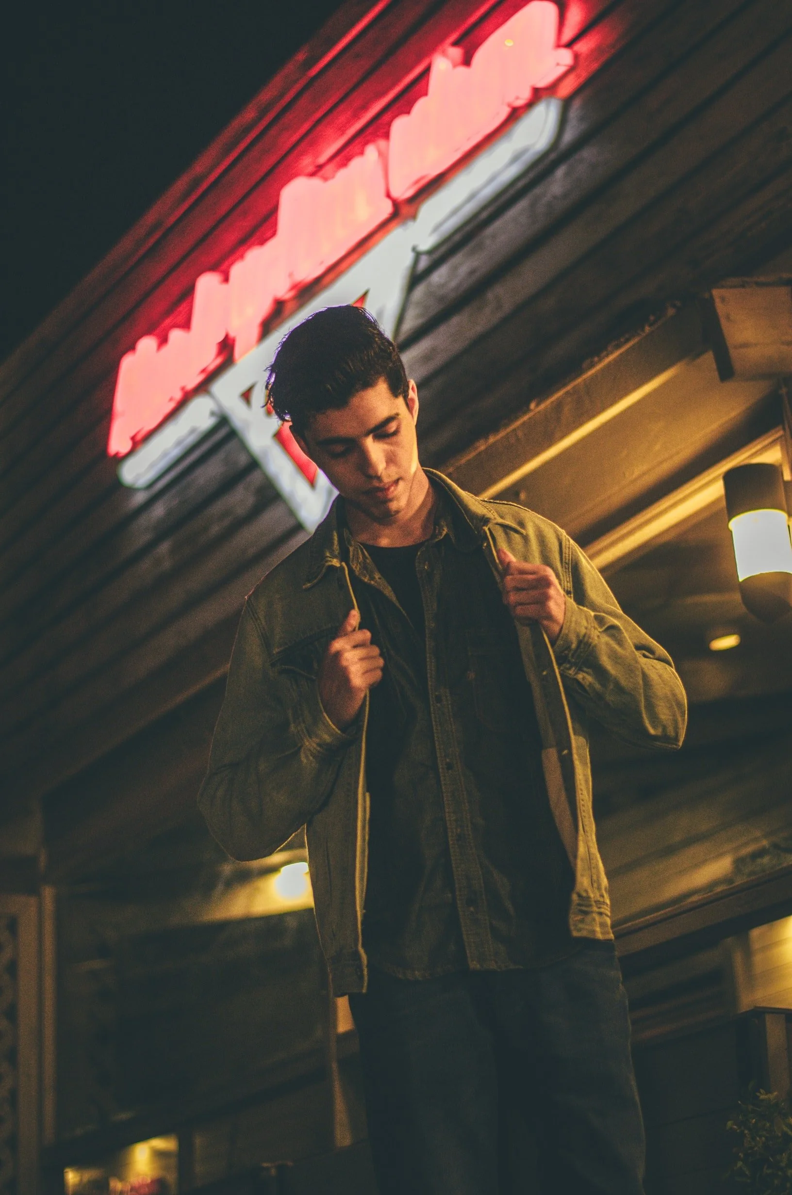 Young man in a denim jacket standing outdoors at night in front of a neon sign.