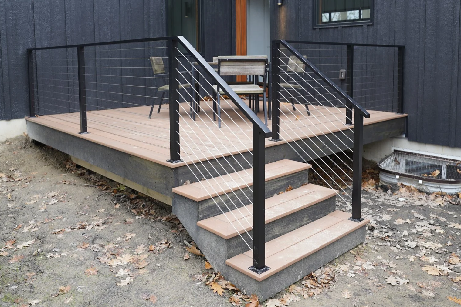 A wooden deck with a black metal and cable railing, accessed by three steps, with outdoor furniture including a table and chairs, and fallen leaves on the ground.