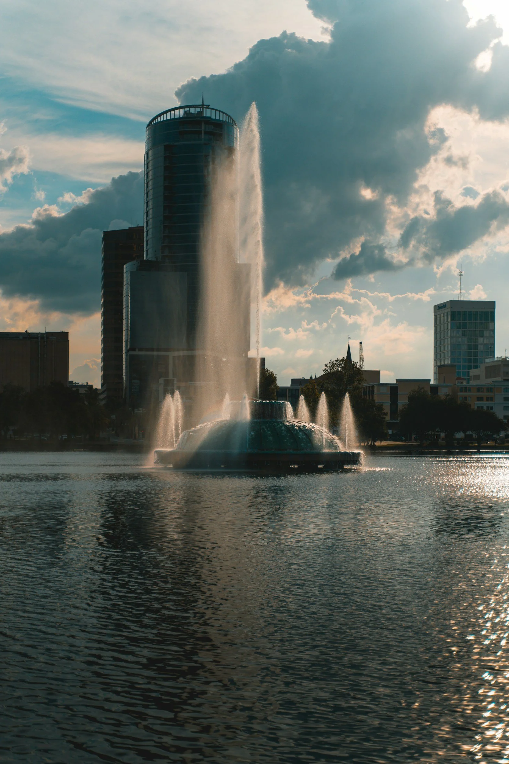 A city skyline with a prominent fountain in the foreground, with water spraying upwards and reflecting on the water's surface, skyscrapers and clouds in the background.