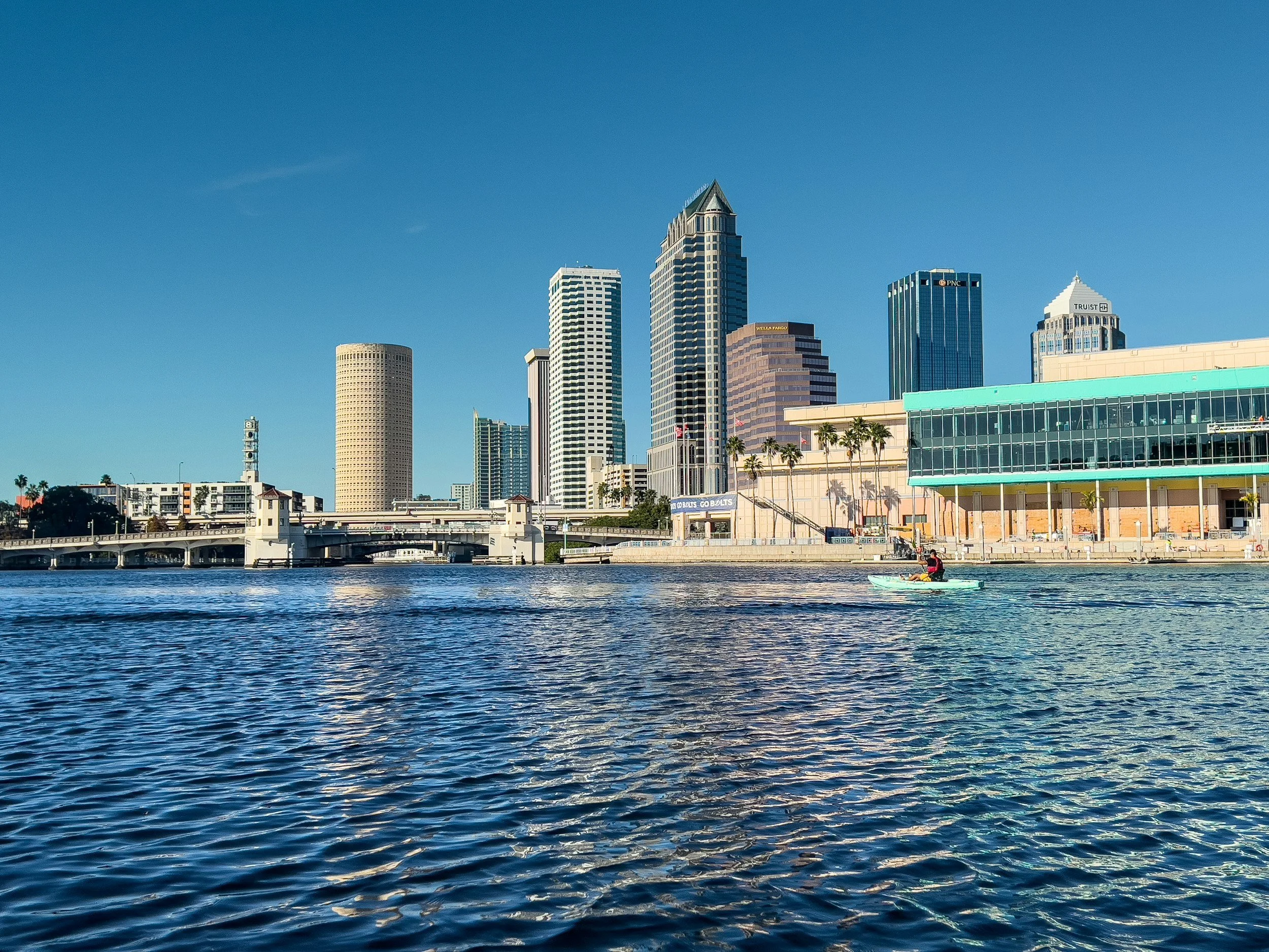 City skyline with tall buildings and a body of water in the foreground, a person kayaking on the water.