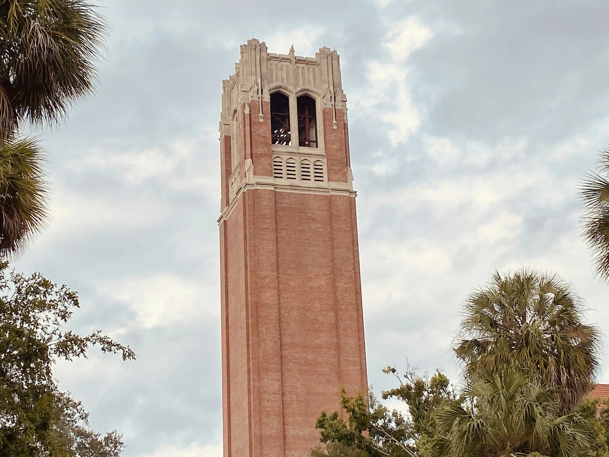 Tall brick clock tower with arched openings at the top, surrounded by trees and under a cloudy sky.