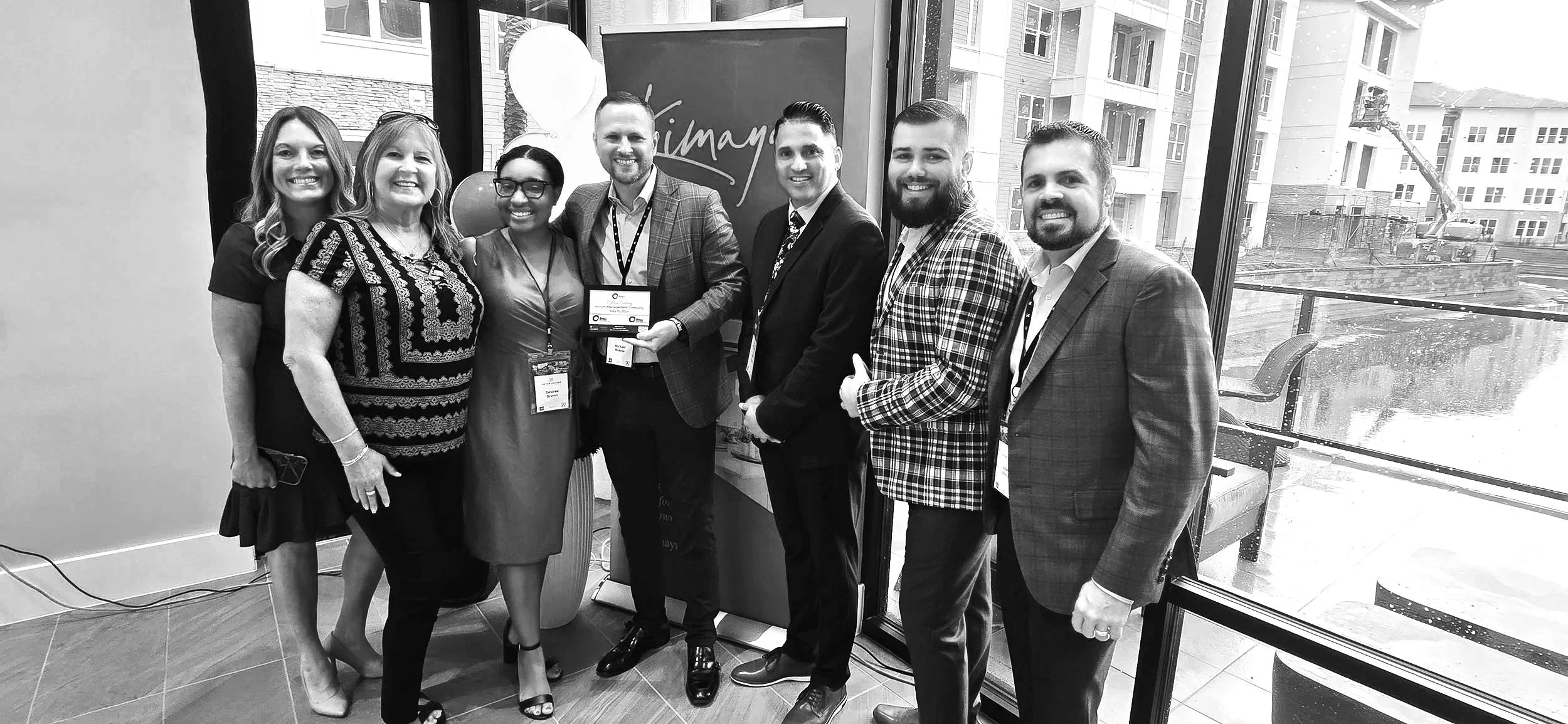 Group of seven professionally dressed people standing inside a building, smiling, with a cityscape visible through the large windows behind them. They are celebrating or attending an event, with one person holding a framed award or certificate.