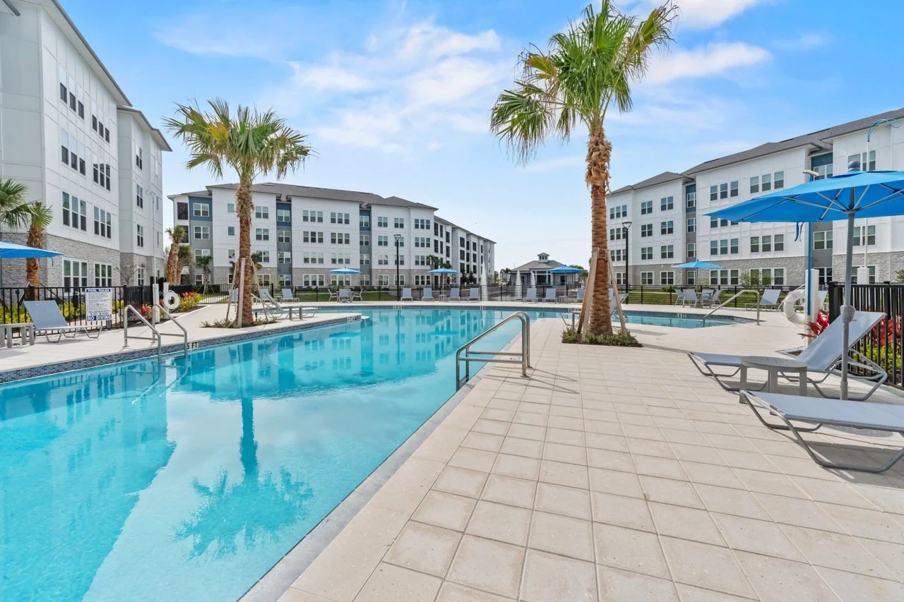 Outdoor swimming pool area surrounded by apartment buildings, with palm trees, lounge chairs, and blue umbrellas under a partly cloudy sky.