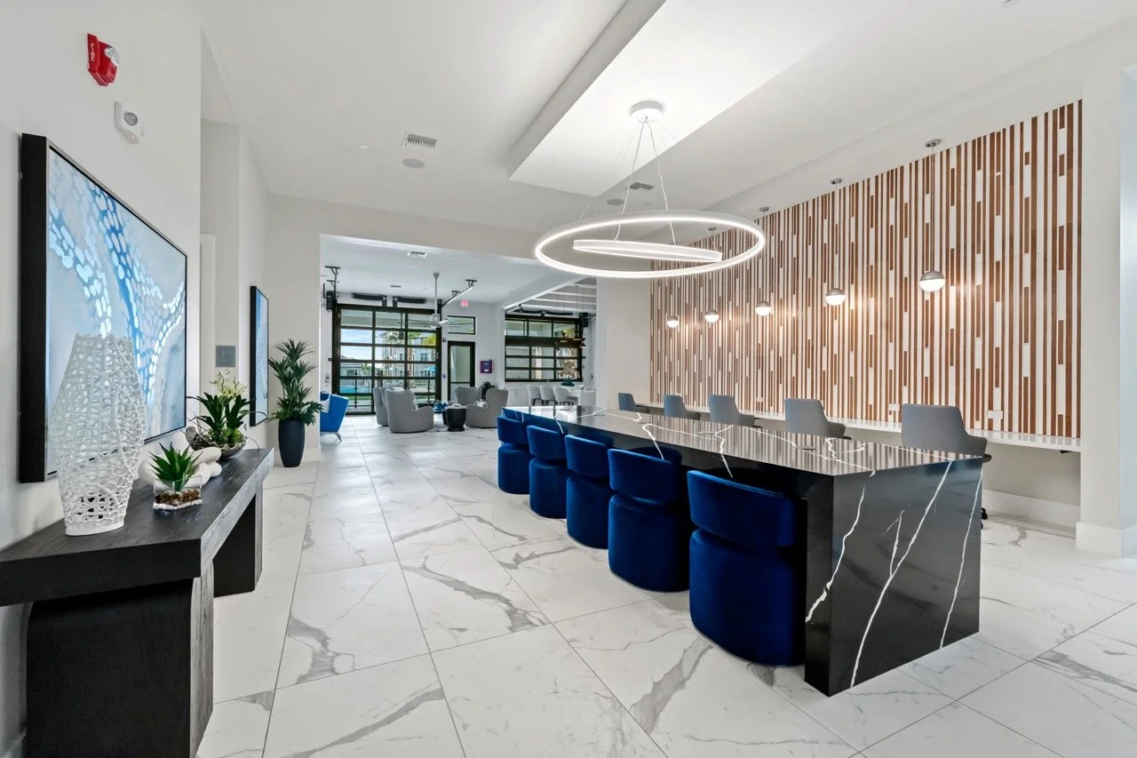 Modern hotel lobby with marble floors, a long black marble table with blue velvet chairs, a wooden accent wall with spherical lights, and a round chandelier. There are seating areas with armchairs and large windows at the back.