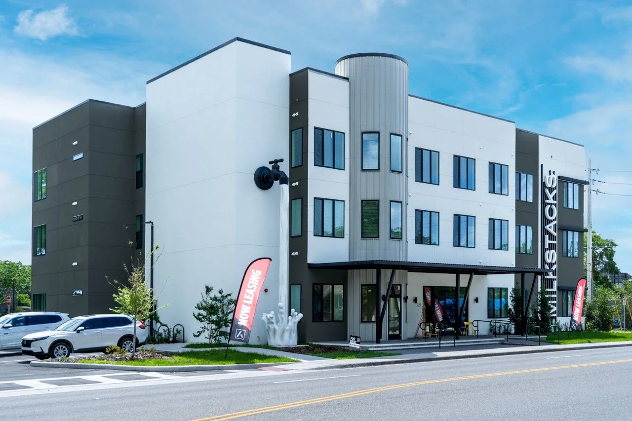 Modern multi-story apartment building with a sign that reads 'VISTA HEIGHTS' and red 'Now Leasing' banners outside. The building has white and dark gray exterior walls, multiple windows, and a covered entrance with a small landscaped area and parked cars in front.