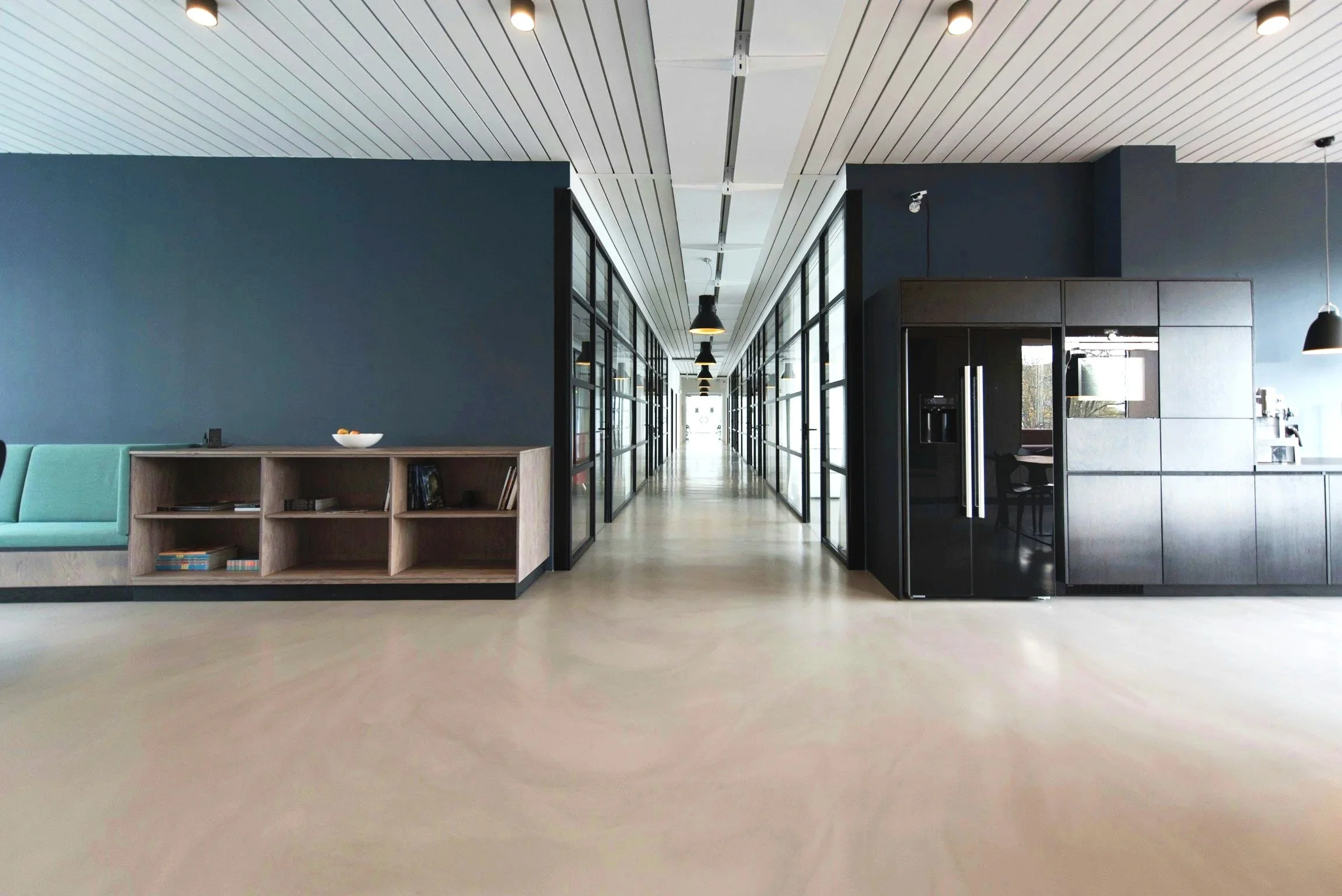 Modern office hallway with glass-walled meeting rooms on both sides, black and gray cabinetry, pendant lights, and a light-colored floor.