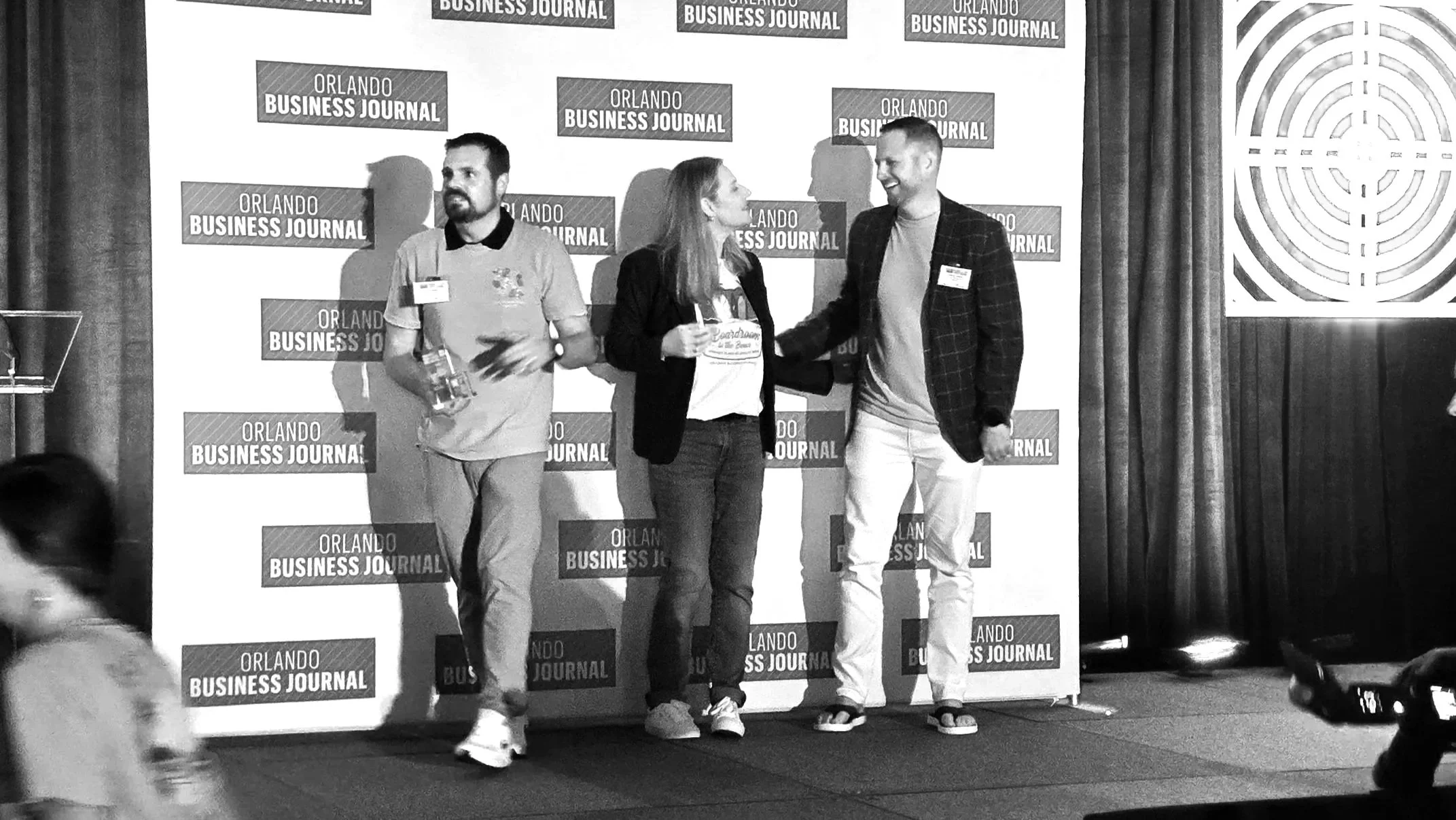 Three people standing on stage at an Orlando Business Journal event, with two men and one woman talking and smiling, backdrop with Orlando Business Journal logos, and curtains on the right side.