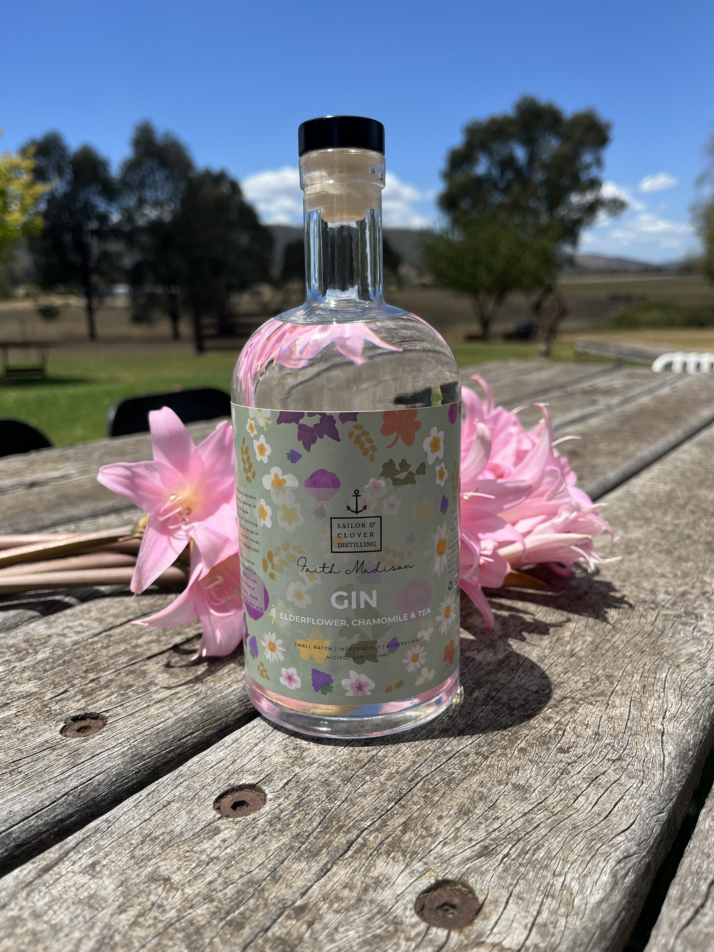 A bottle of Sailor & Clover Distilling gin with pink flowers beside it on a weathered wooden outdoor table, with a rural landscape and clear blue sky in the background.