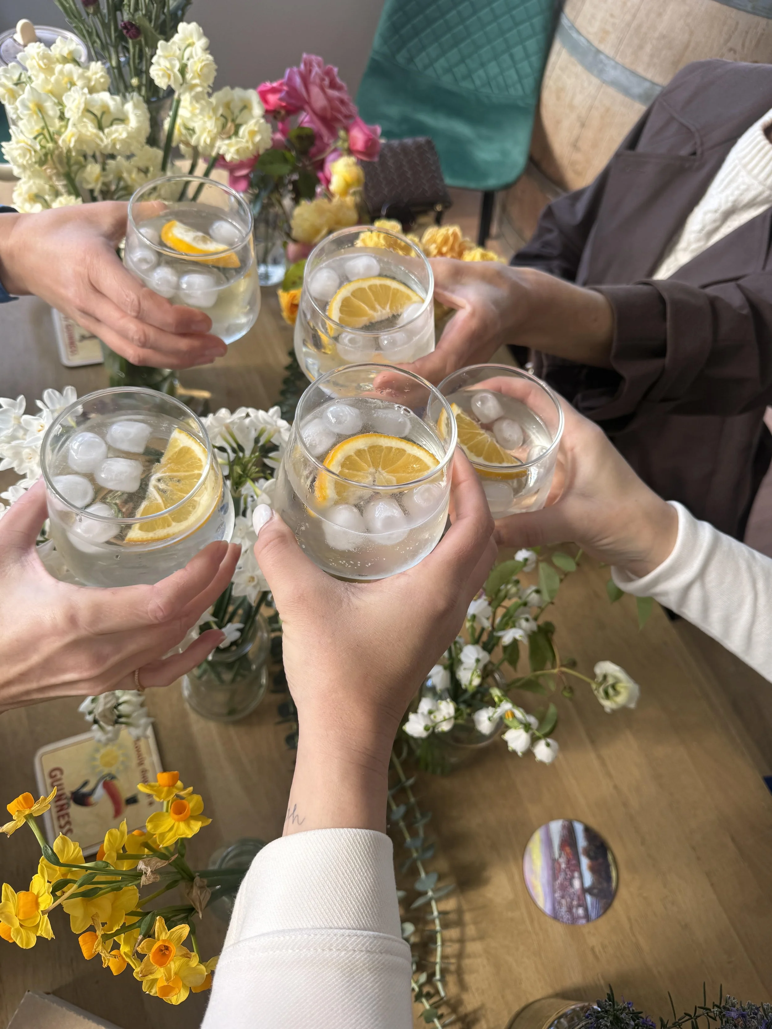 Three people raising glasses filled with ice, lemon slices, and clear beverage at a floral-themed gathering.