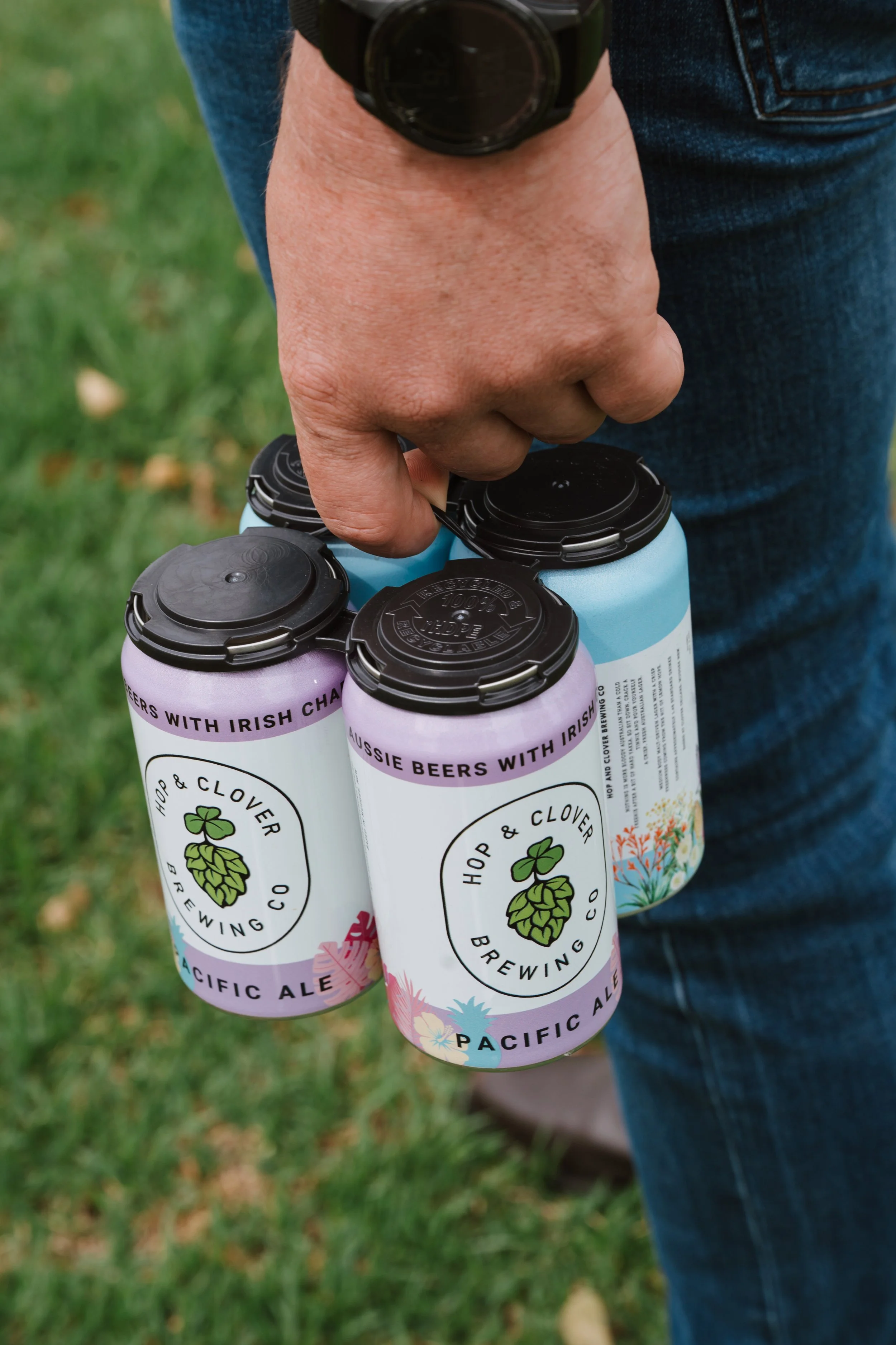 A person holding four cans of beer outdoors on green grass, with denim jeans visible.