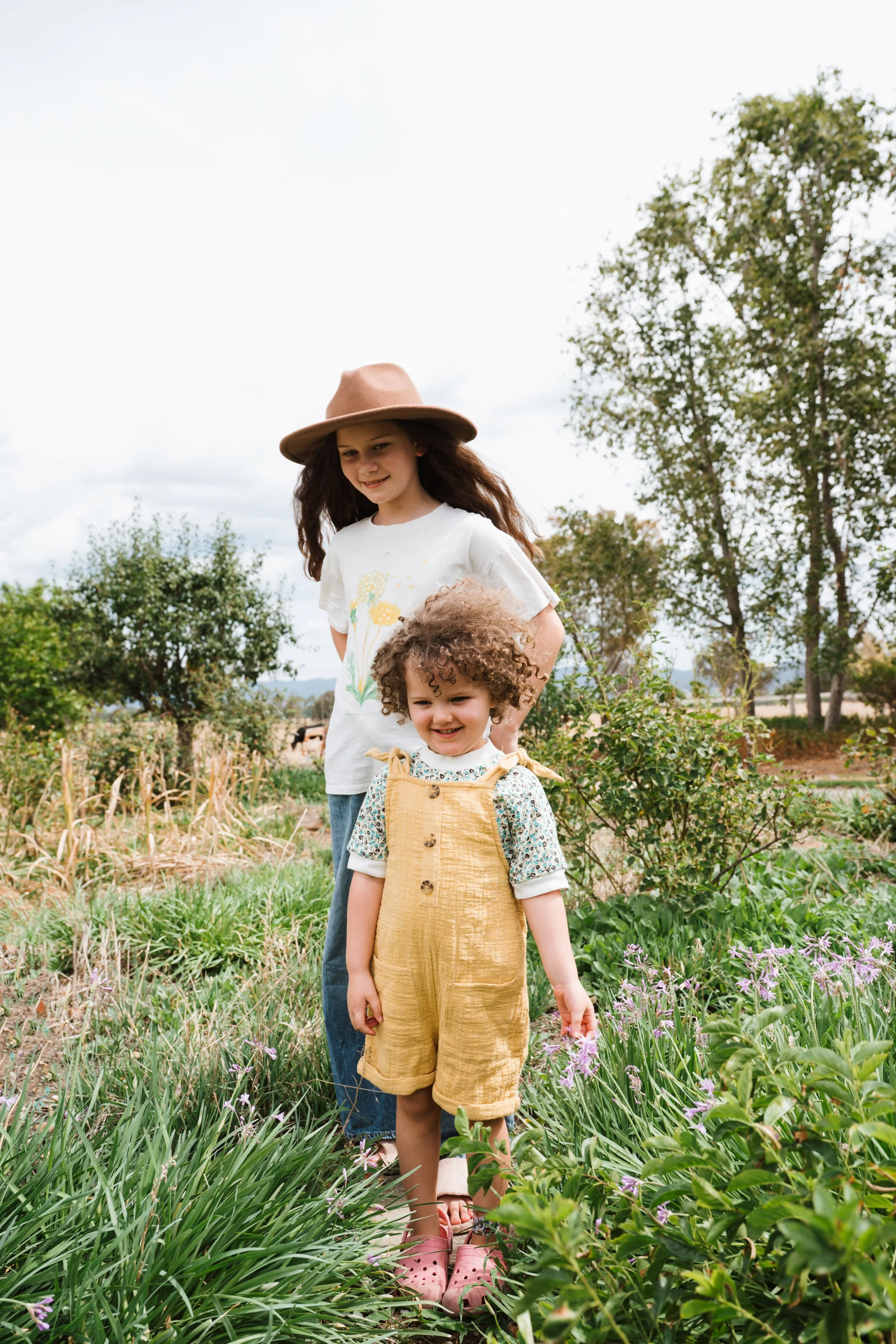 Two young girls, one older with long curly hair wearing a wide-brimmed hat, white t-shirt, and jeans, and the younger with short curly hair wearing a tan overalls and floral shirt, standing in a garden with bushes, trees, and purple flowers, overcast sky.