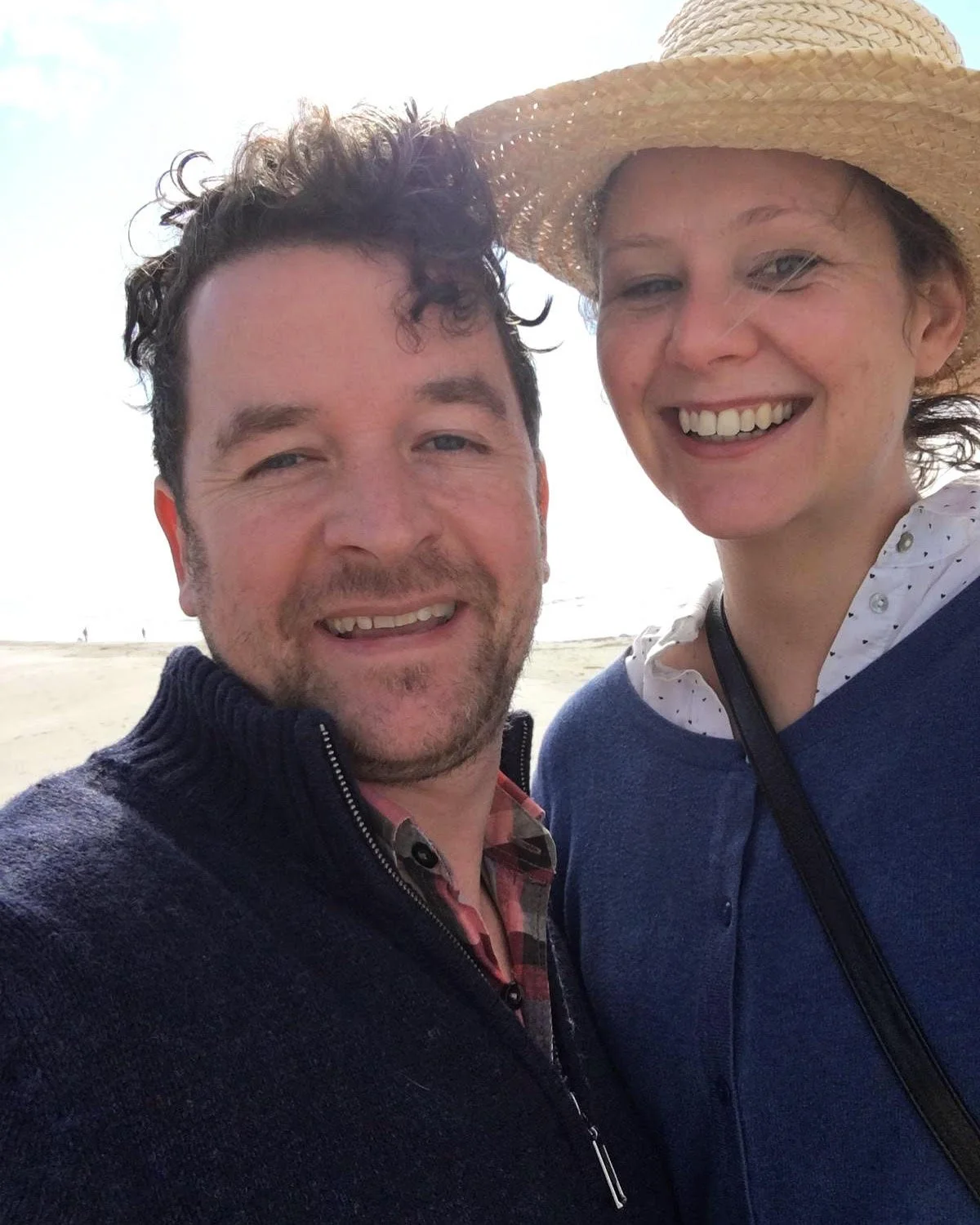 A close-up photo of a smiling man and woman on the beach, with the woman wearing a large straw hat.