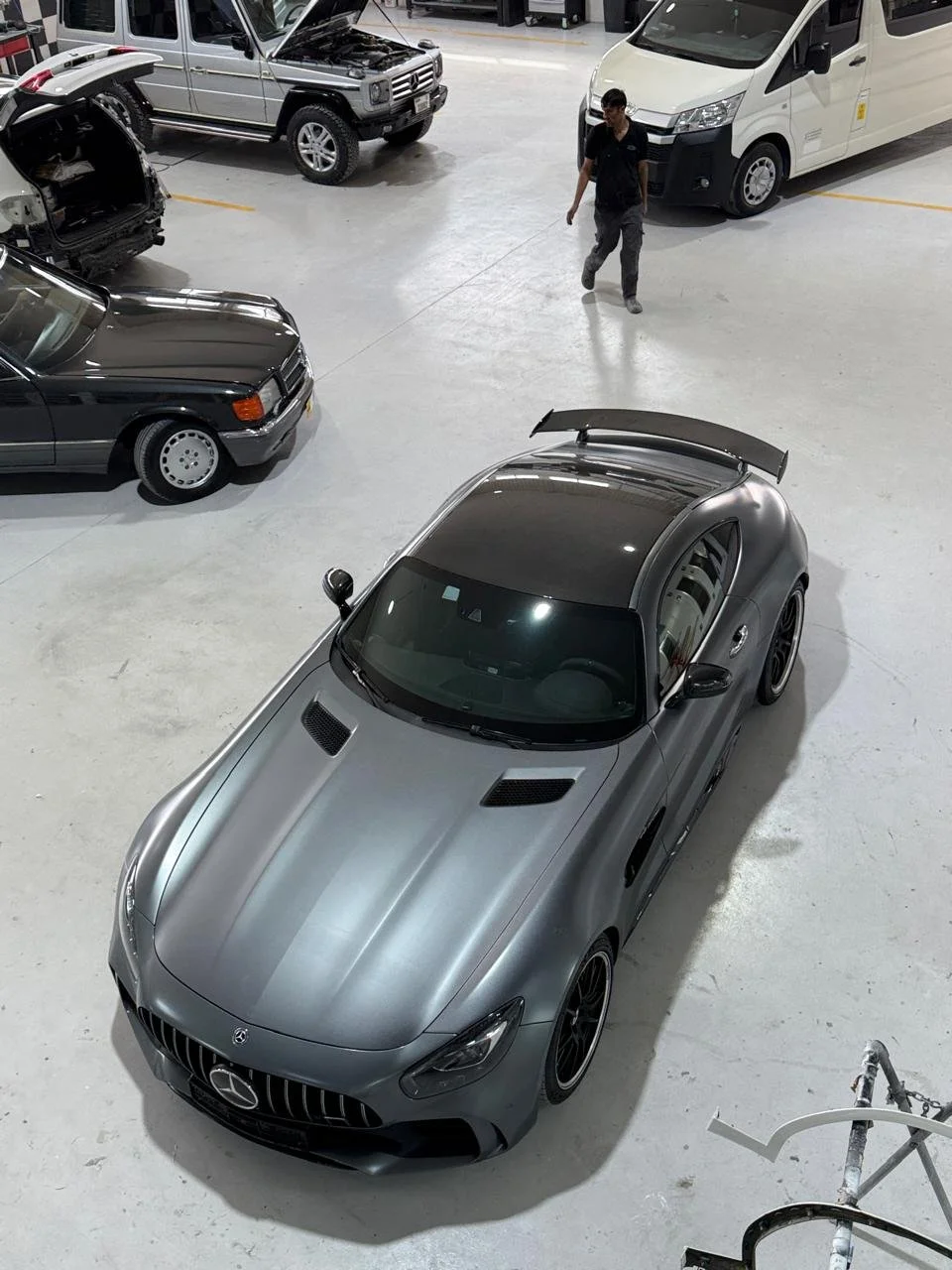 A silver Mercedes-Benz sports car with a large rear wing is parked inside Star Centre car body shop, surrounded by other vehicles including a black classic car, a grey SUV, and a white van. A Star Centre employee is walking nearby.