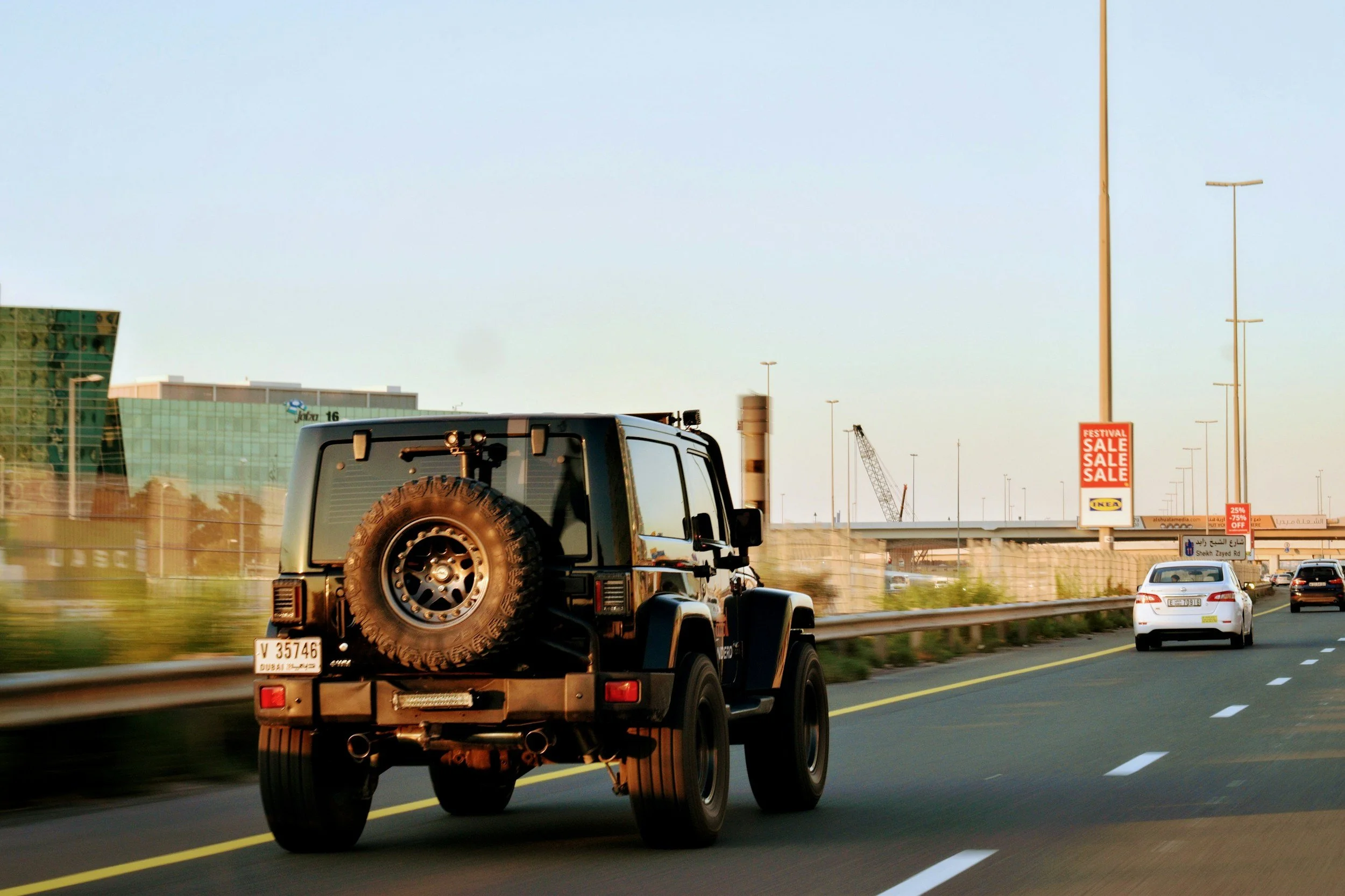 A black Jeep with a spare tire on the back driving on a multi-lane highway in Dubai, with buildings, street lamps, and billboards including a sale sign visible in the background.