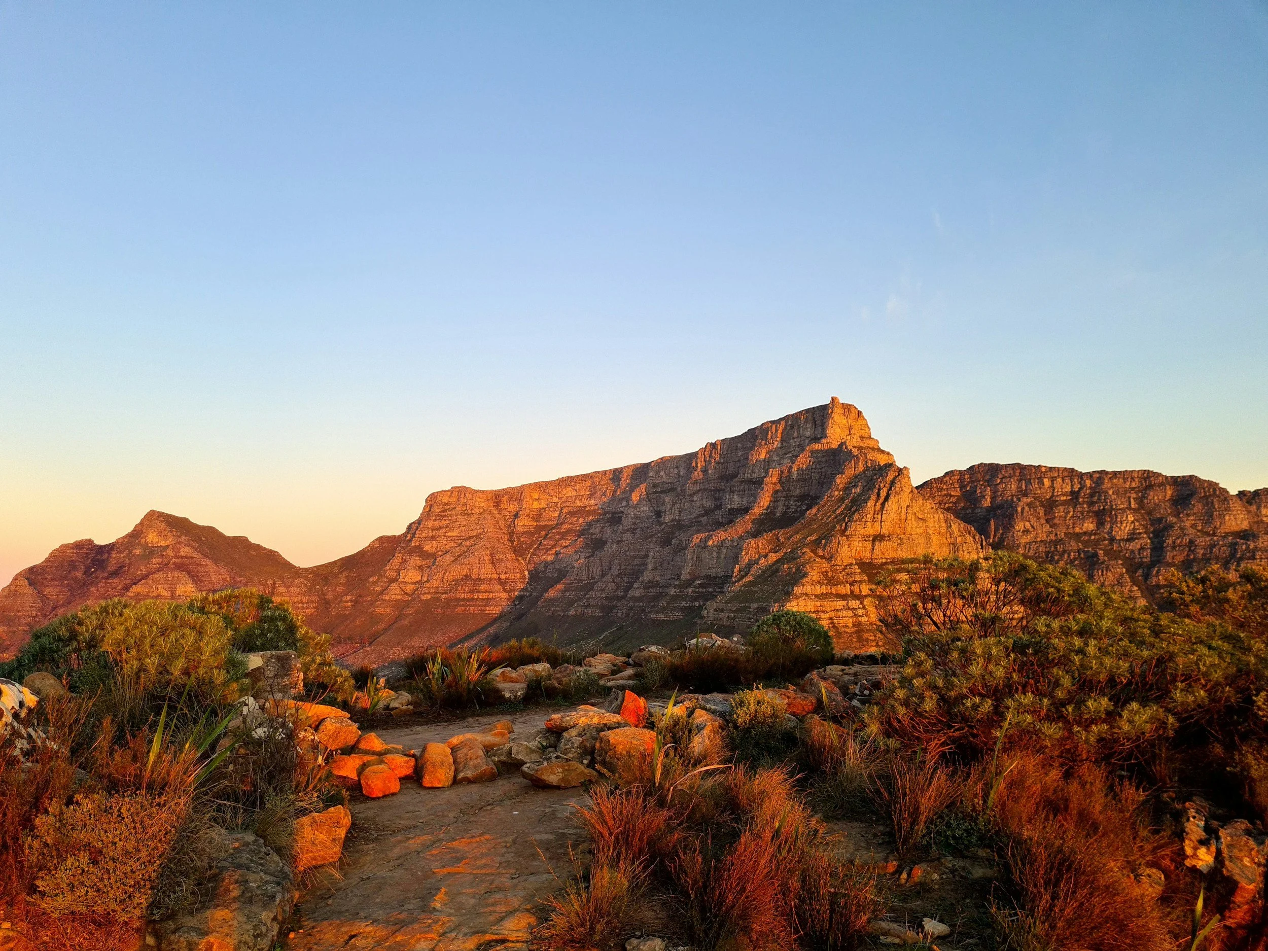 Sunset over rocky mountain terrain with dry shrubs and plants in foreground.