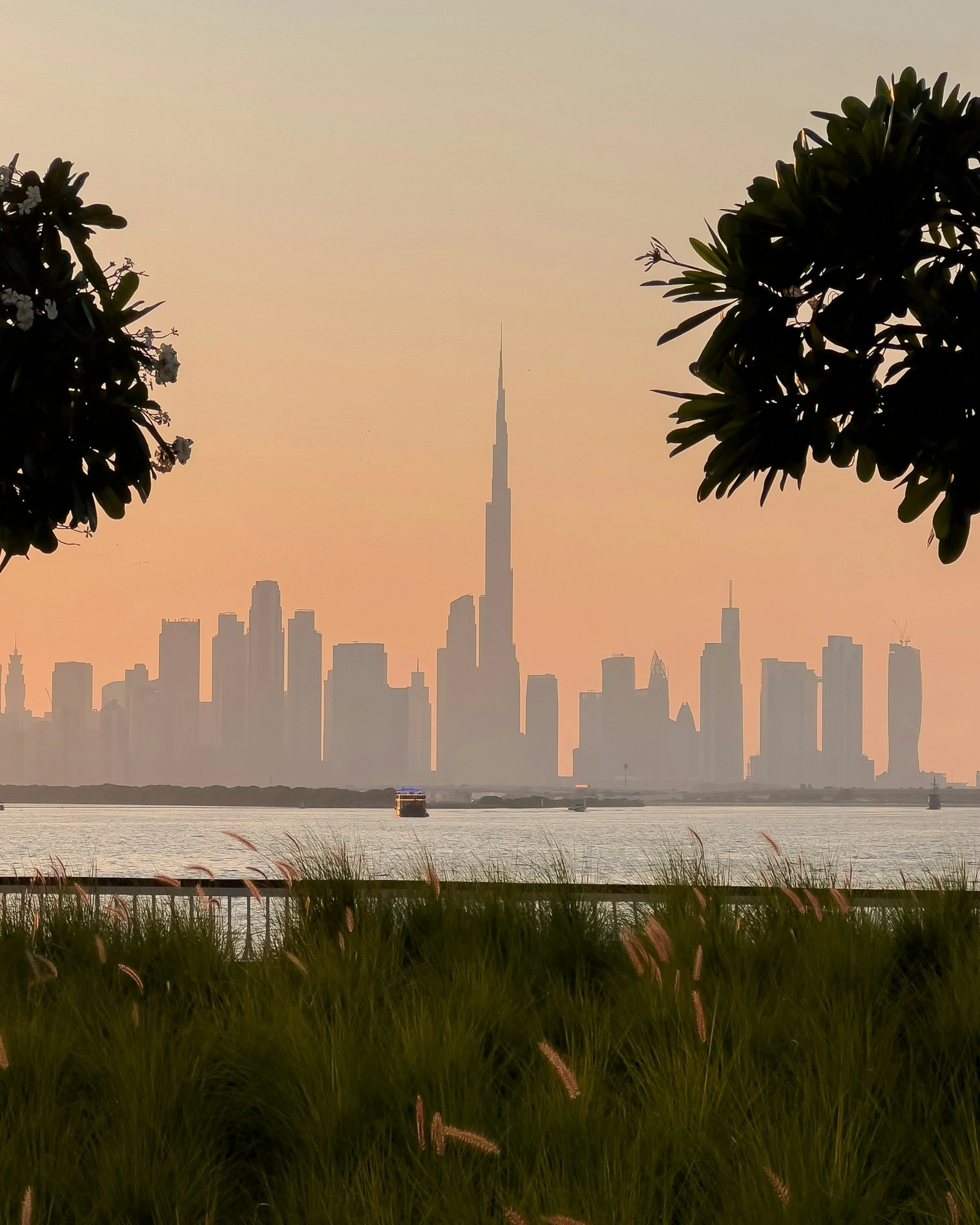 Silhouette of a city skyline, including the Burj Khalifa, at sunset with water in the foreground and trees on either side.